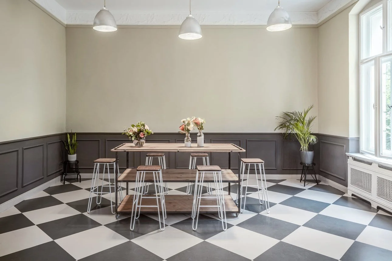 Sala da pranzo luminosa con pareti bianche e pannelli grigi, tavolo in legno con sedia e panca, vasi con fiori e piante verdi, grande finestra, pavimento a scacchi bianco e nero.