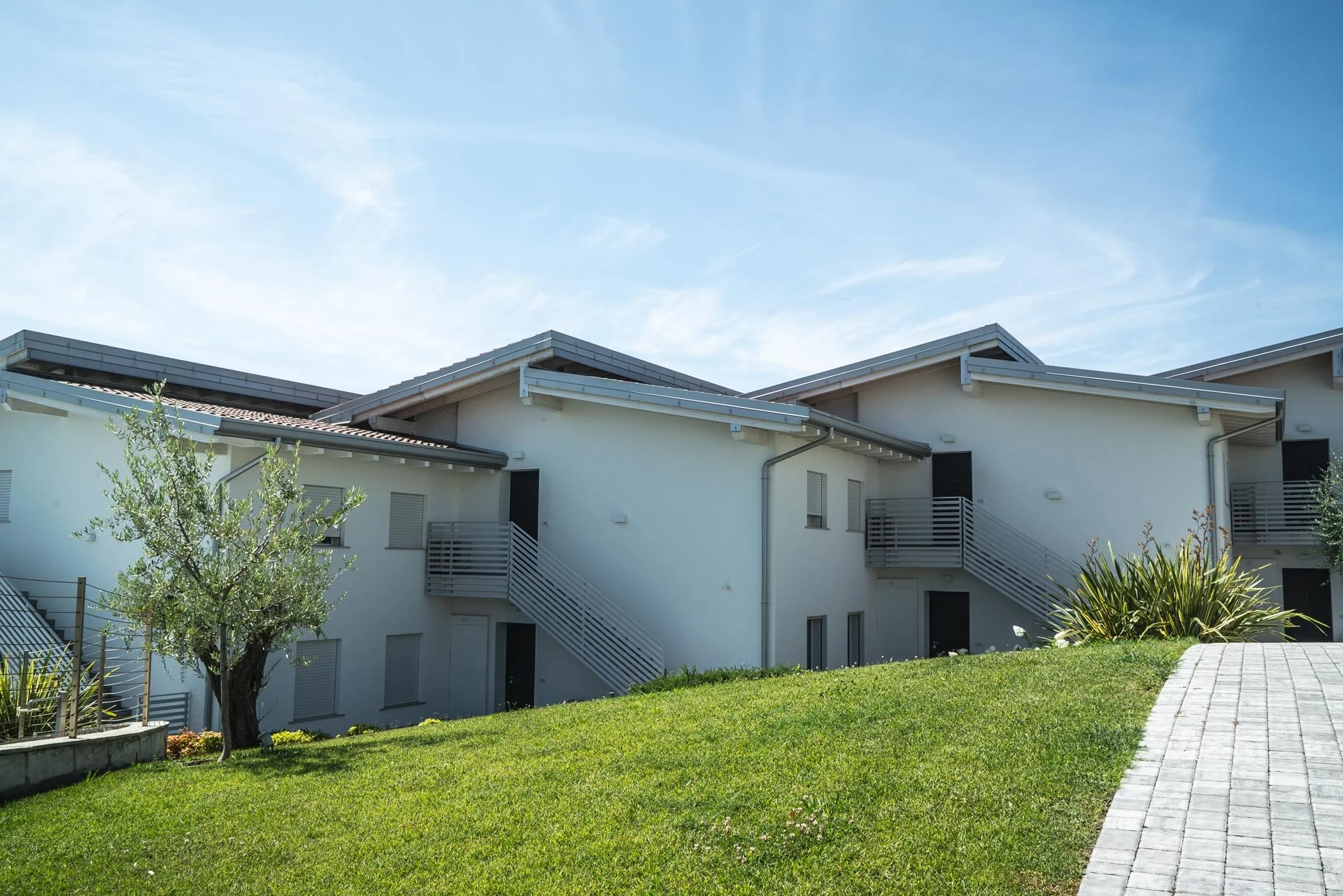Complesso residenziale con case bianche, tetti a falda grigi e scale esterne, circondato da prato verde e albero di fronte, sotto un cielo azzurro.