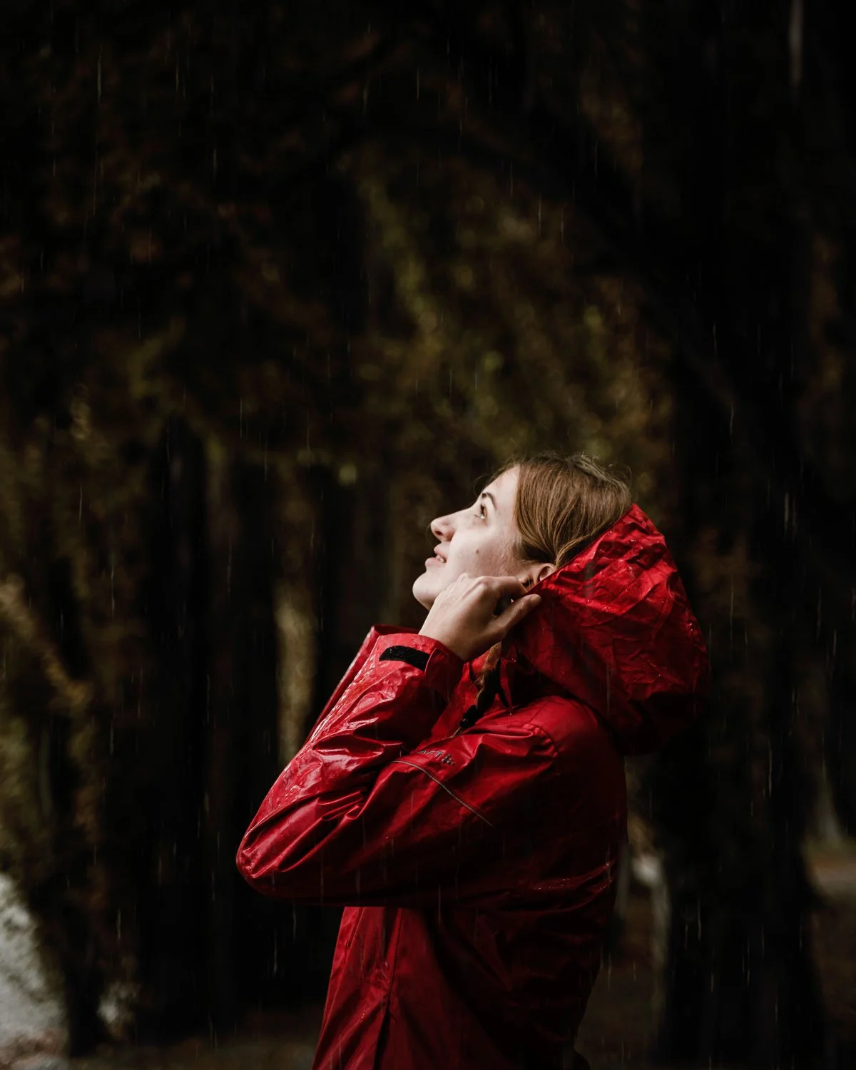 A woman in a red waterproof jacket, holding the hood, standing outdoors in the rain, looking up with a happy expression.