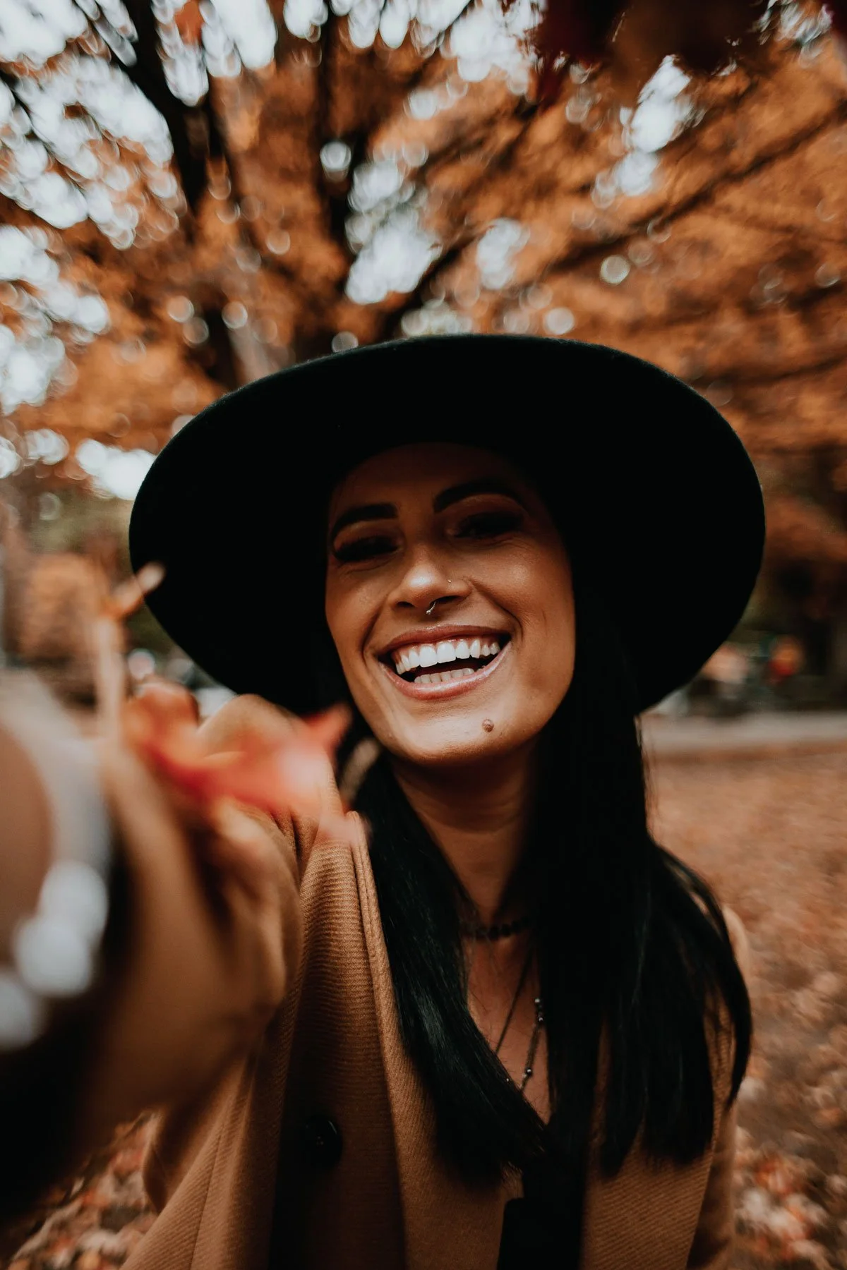A woman smiling and reaching toward the camera, wearing a large black hat and a tan coat, with autumn leaves and blurred trees in the background.