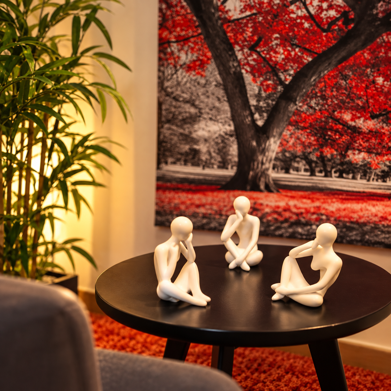 Figurine di persone sconosciute sedute intorno a un tavolo nero, con un grande quadro di un albero autunnale sullo sfondo e una pianta verde sulla sinistra.