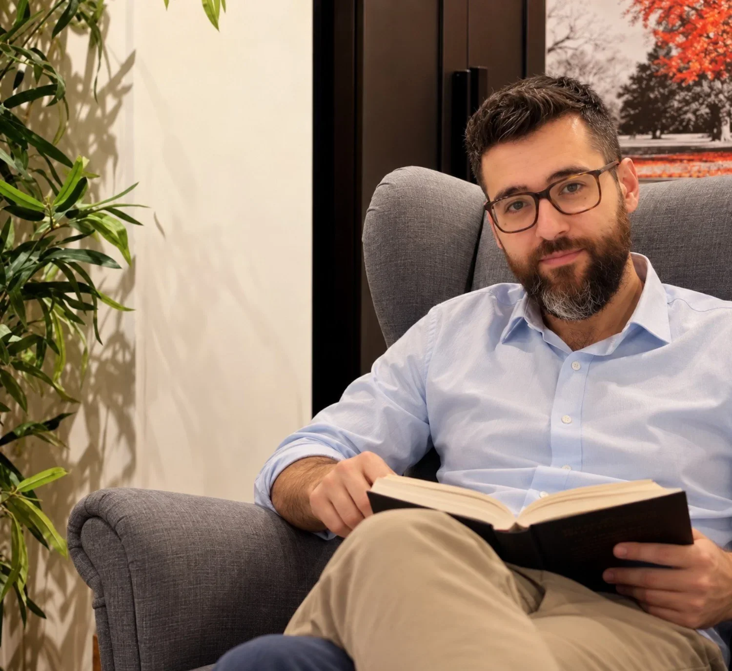Uomo con barba e occhiali seduto su una poltrona, che legge un libro.