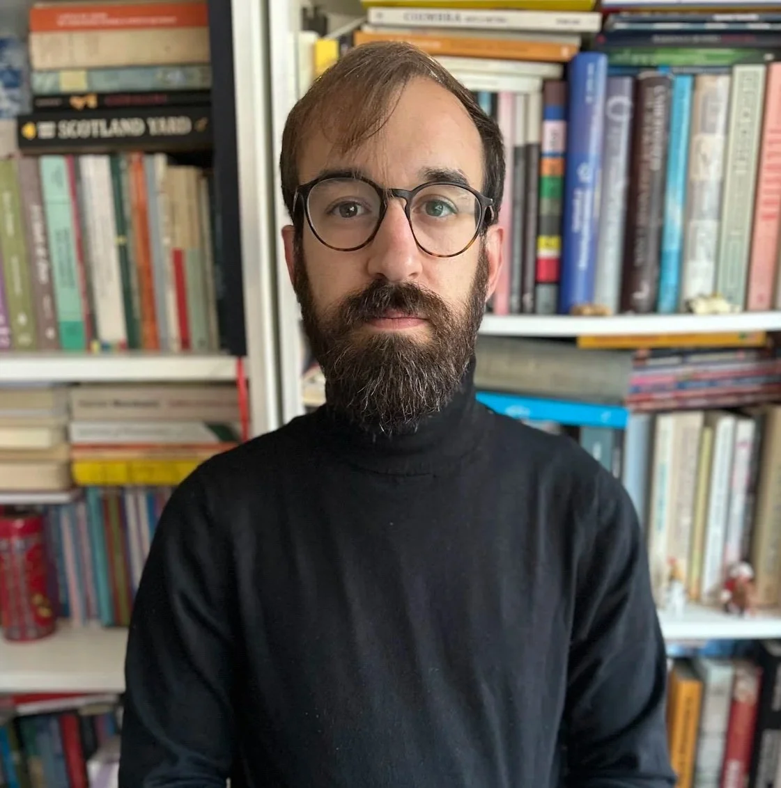 Homem com barba, óculos, cabelo castanho claro e camiseta preta, posando na frente de uma estante cheia de livros