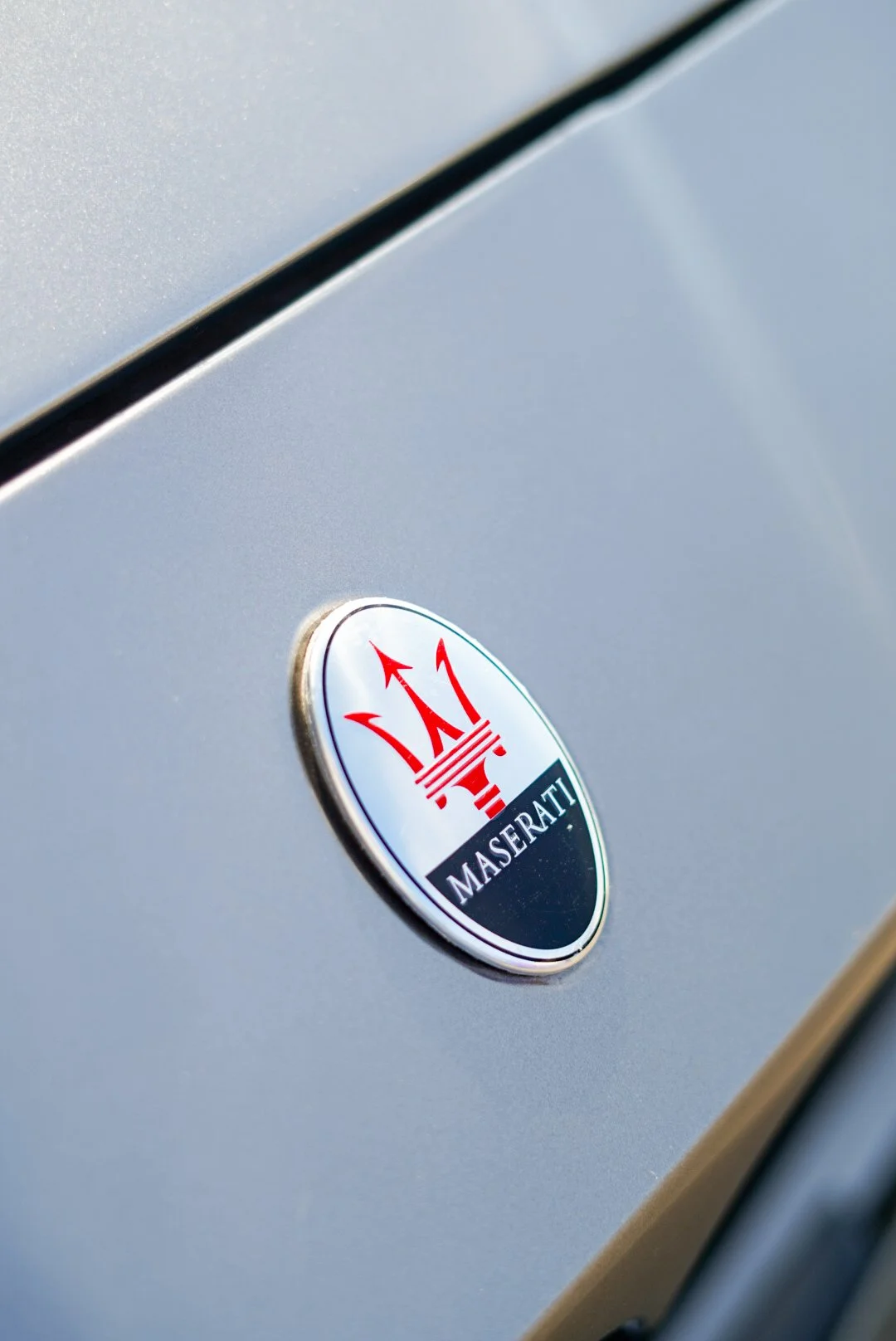 Gros plan sur l’emblématique logo Maserati, un trident rouge et bleu sur carrosserie argentée. Photographie détaillée mettant en valeur le design premium et l'élégance de la marque italienne de luxe. Capturée par Thomas Lotito.