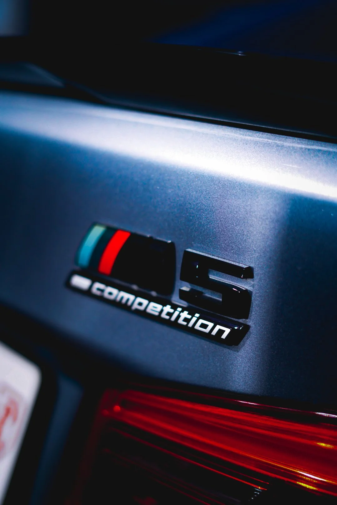 Gros plan sur l’emblème BMW M5 Competition, affichant le badge iconique sur une carrosserie métallisée. Capture détaillée mettant en avant l’ADN sportif et hautes performances du modèle. Photographie réalisée par Thomas Lotito.