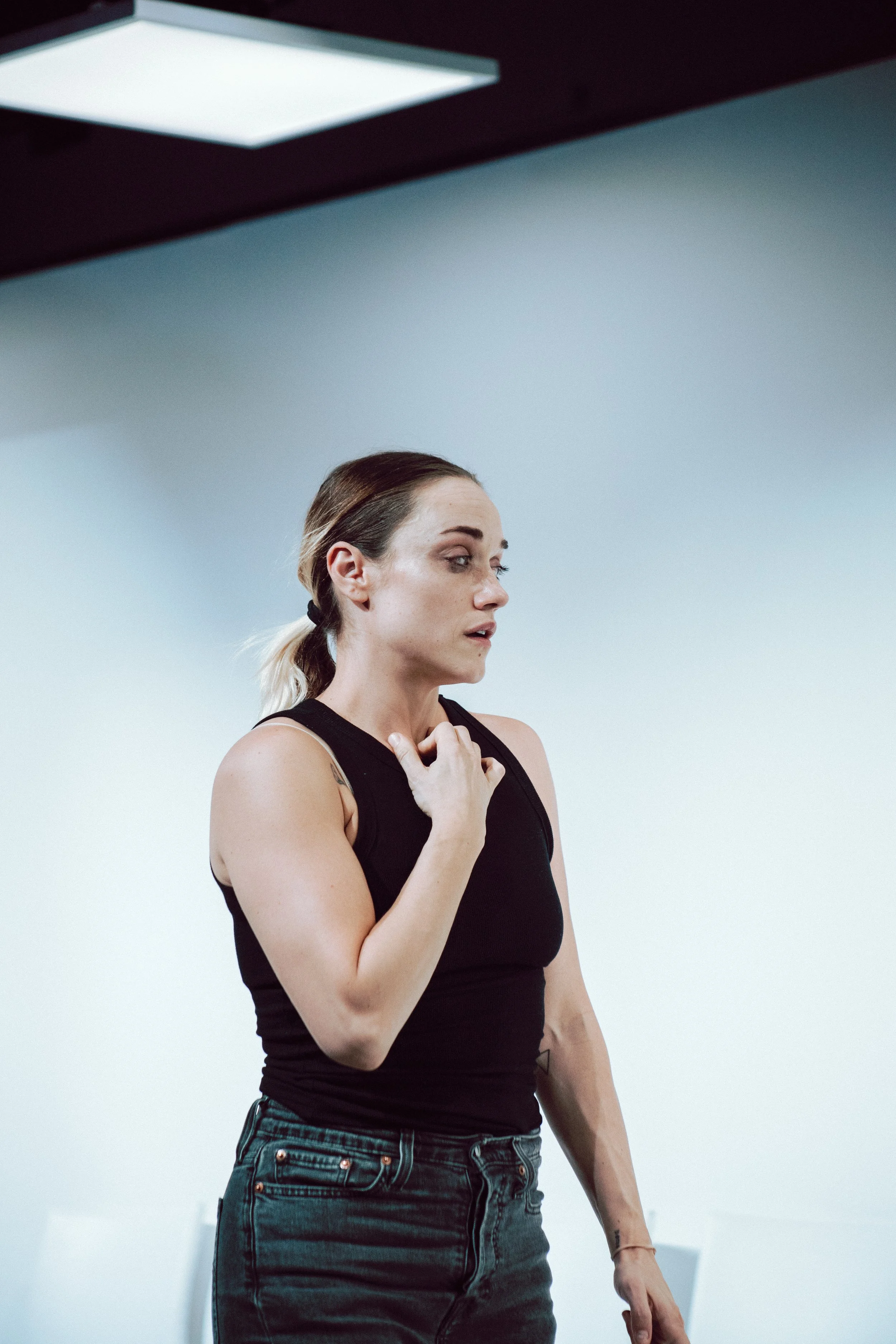 A woman with blonde hair tied back, wearing a black sleeveless top and dark jeans, stands in a room with plain white walls and blue accents, holding her hand to her chest and looking to the side.