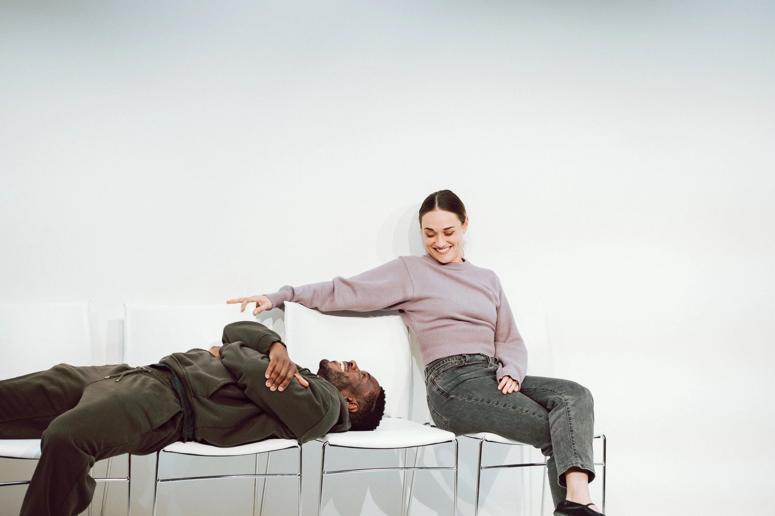 A woman smiling and sitting on a chair next to a man lying down with his eyes closed, all in a bright, minimal room.