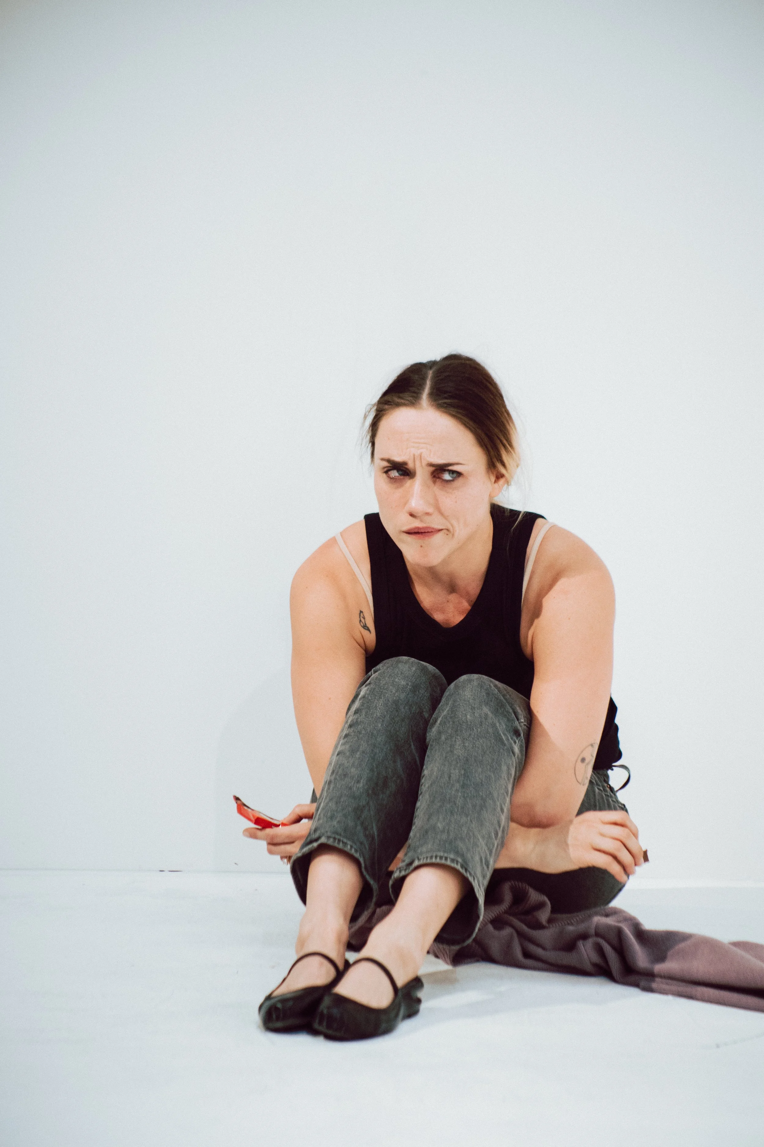 A woman with brown hair and tattoos, sitting on the floor with her knees up and holding a red phone, looks confused or questioning, against a plain white background.