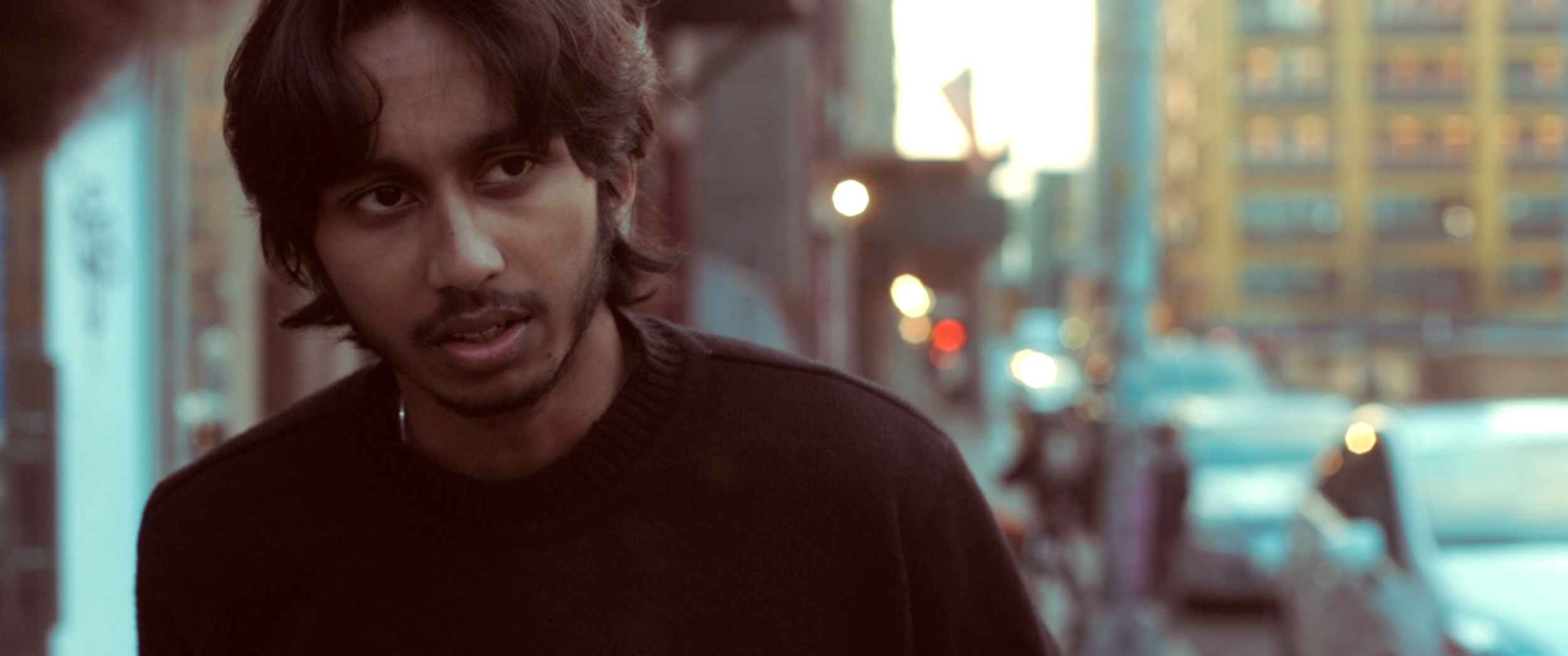 A young man with medium-length wavy hair and a beard wearing a black sweater, speaking to someone, in an urban street setting during evening or dusk, with blurred city lights and buildings in the background.