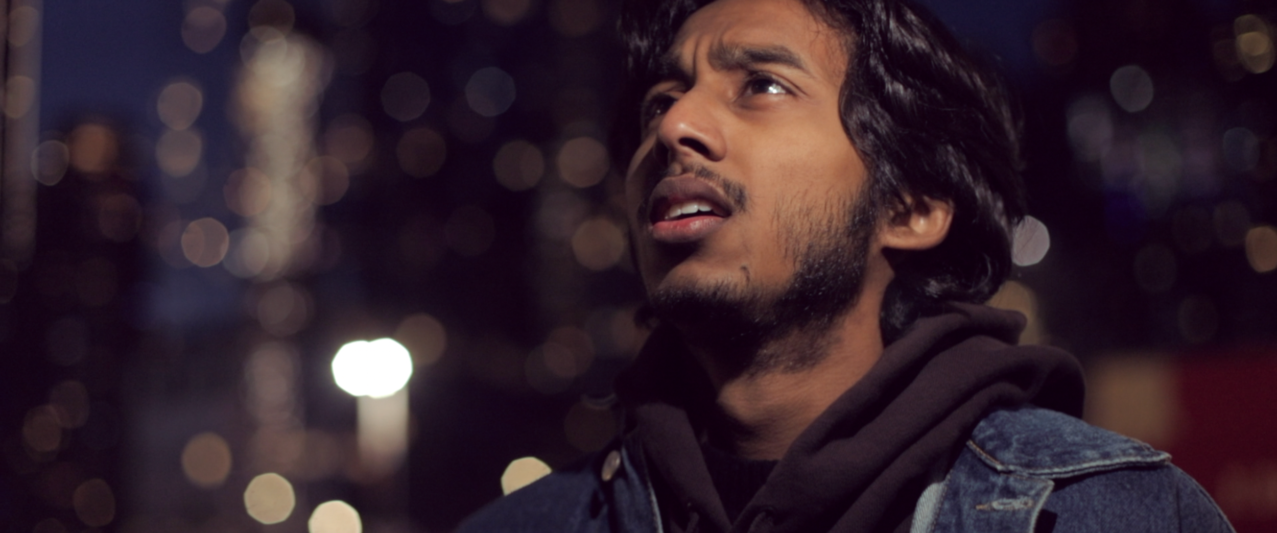 A young man with long dark hair and facial hair looking upward against a city nightscape with bokeh lights.