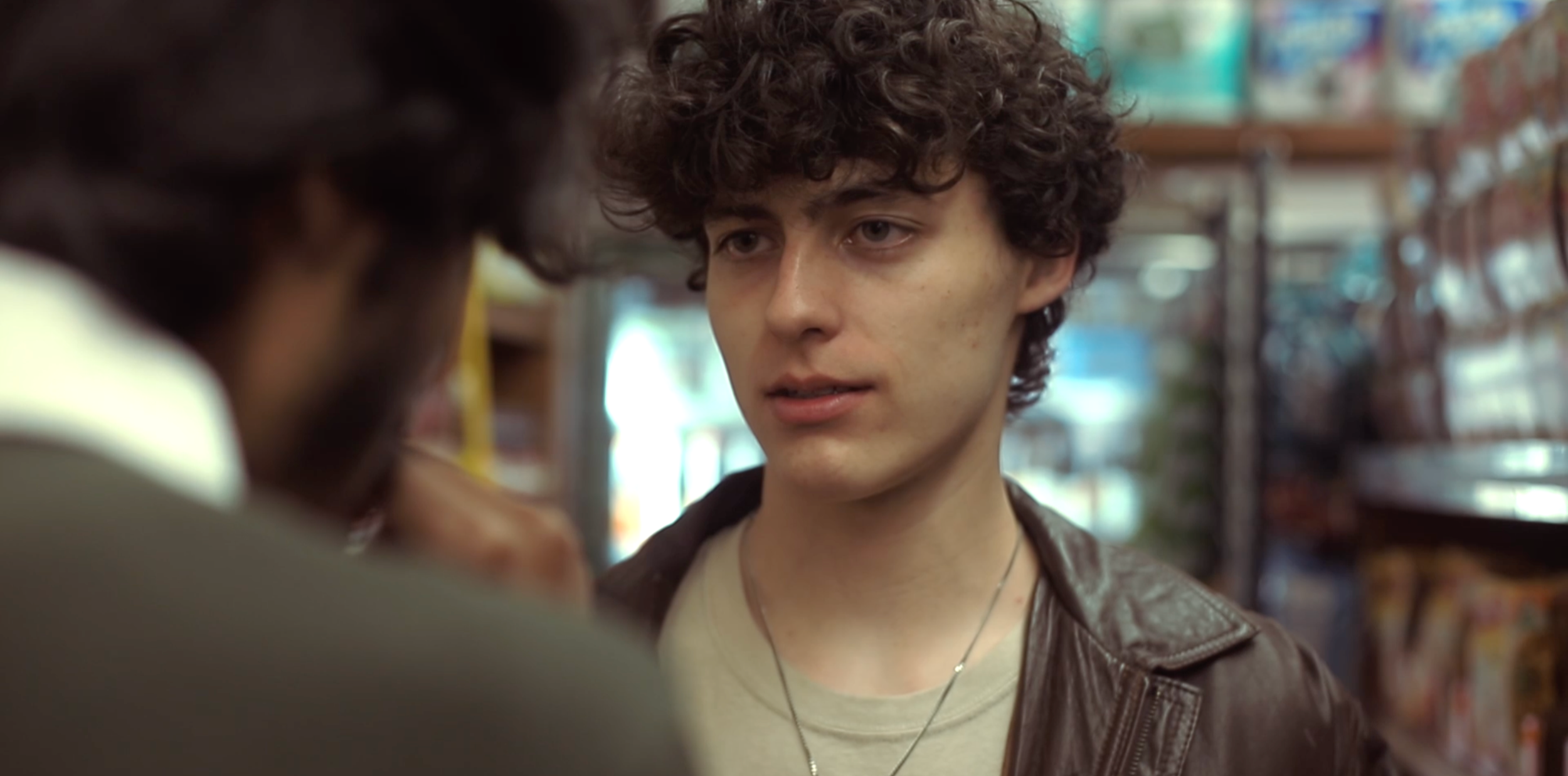 A young man with curly hair and a leather jacket looks at another person in a store aisle.