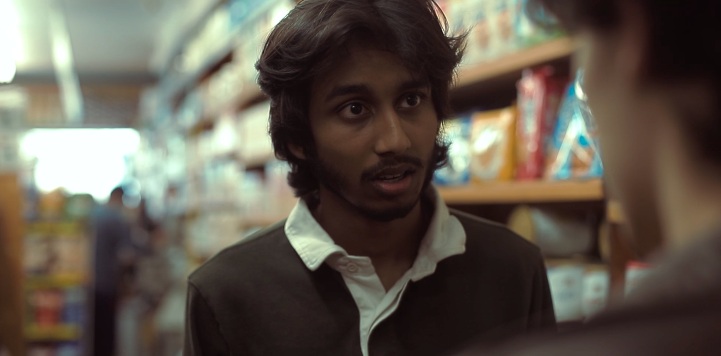 A young man with dark, wavy hair and a goatee looking at something in a store aisle, with shelves stocked with snack bags in the background.