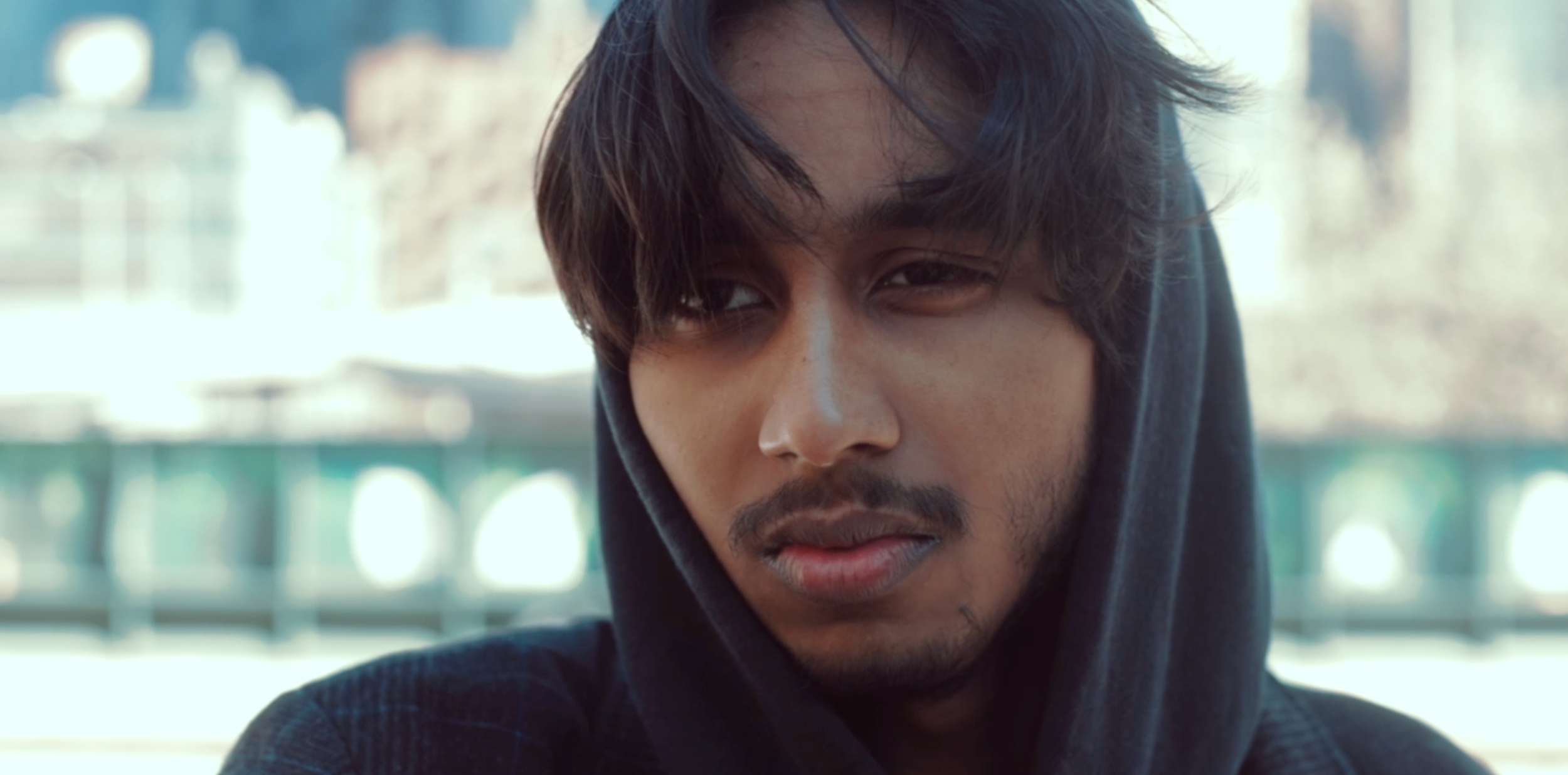 Close-up of a young man with dark hair, a mustache, and facial hair, wearing a black hoodie, looking slightly away in an urban outdoor setting with blurred buildings and trees in the background.
