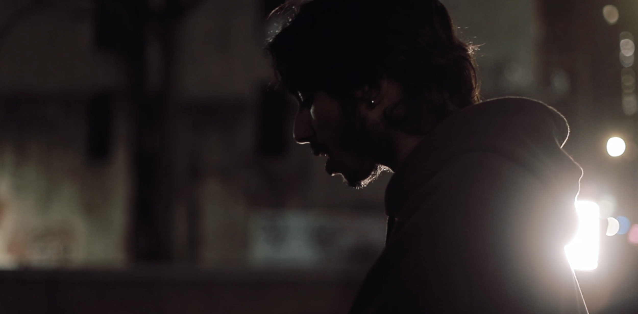 A silhouette of a man with shoulder-length hair and glasses, looking downward, illuminated by backlighting in a dark setting.