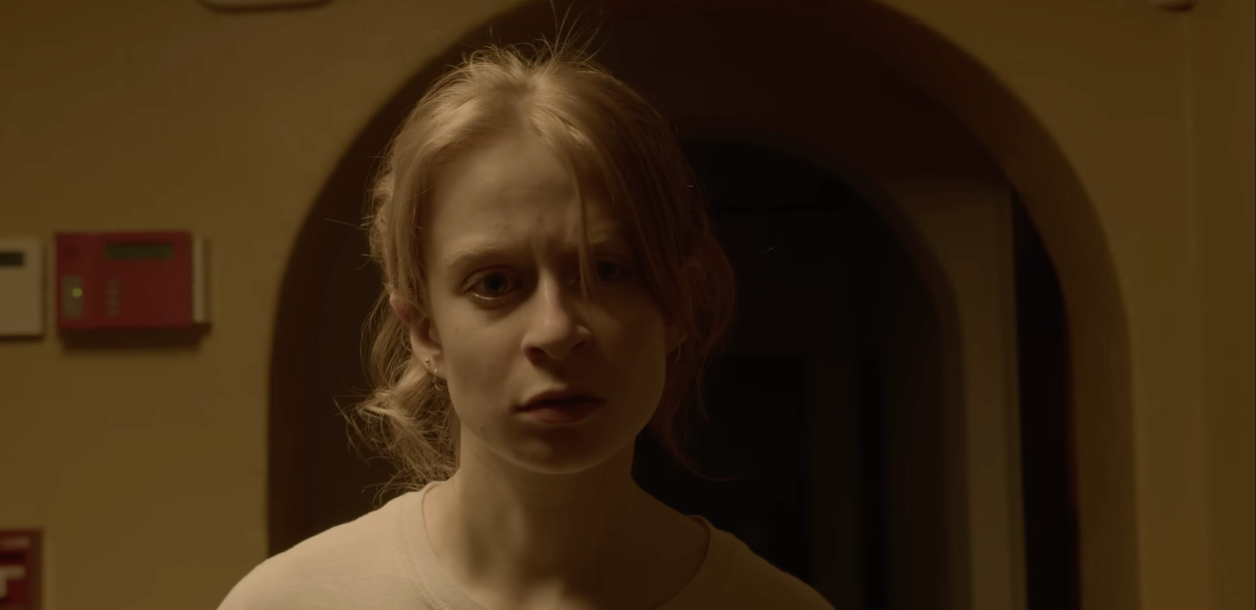 A woman with reddish hair and pale skin standing in front of a dark arched doorway, with a beige wall behind her and a red fire alarm box on the wall.