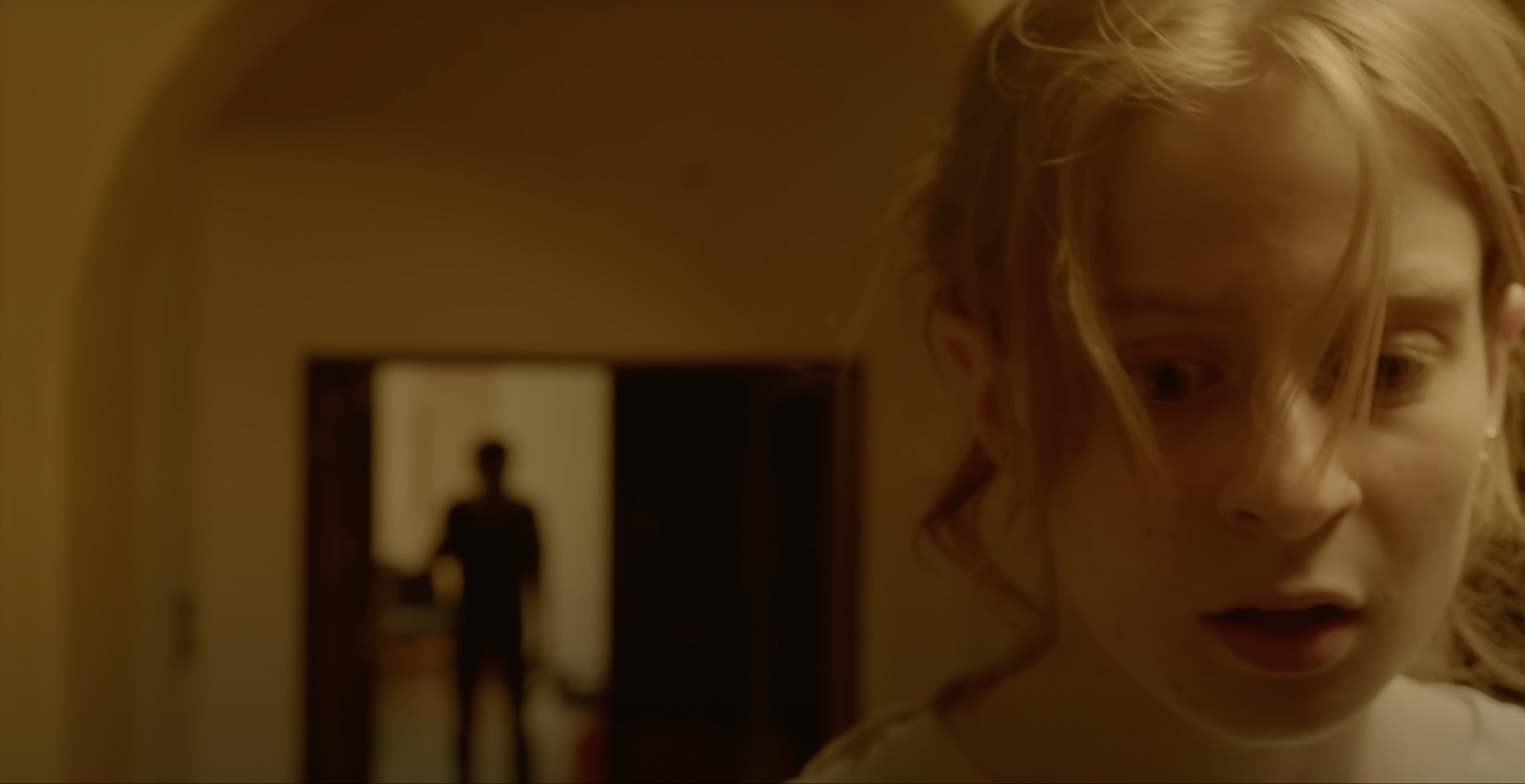Close-up of a young girl with curly blonde hair and a worried expression, with a blurred figure of a person standing in a doorway in the background.