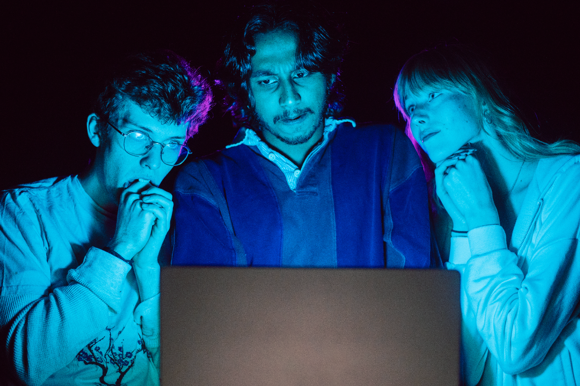 Three young people, two women and one man, sitting together in front of a laptop with blue and purple lighting, showing expressions of surprise or concentration.