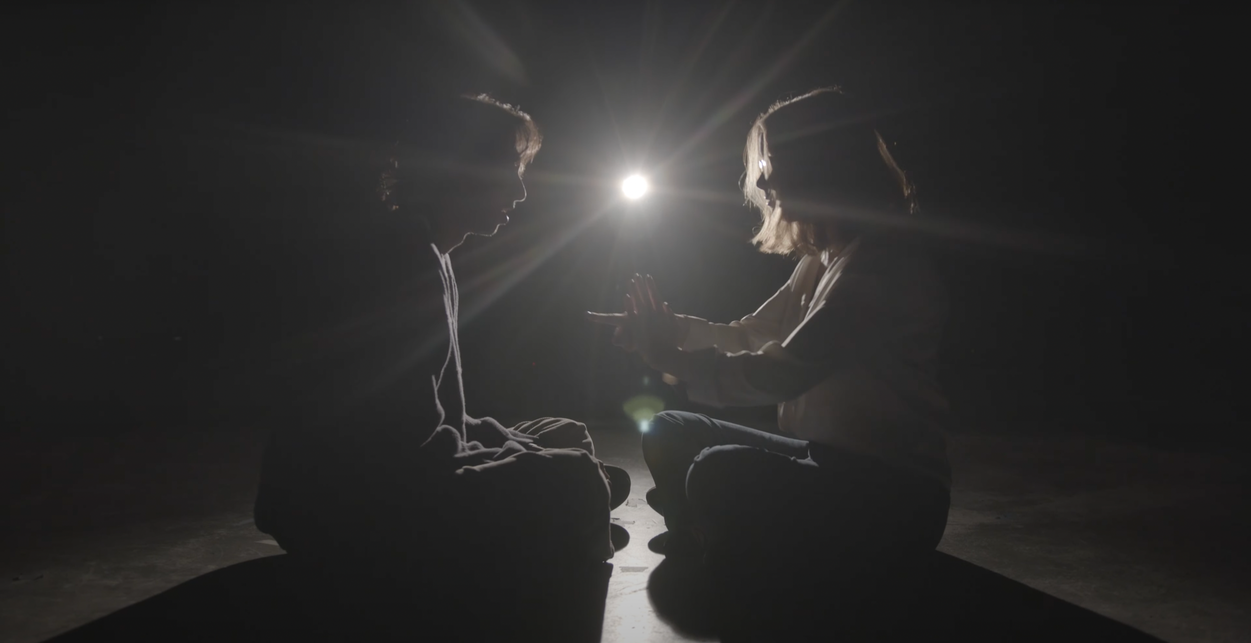 Two children sitting cross-legged in a dark room with a bright light behind them, touching hands in front of their faces.