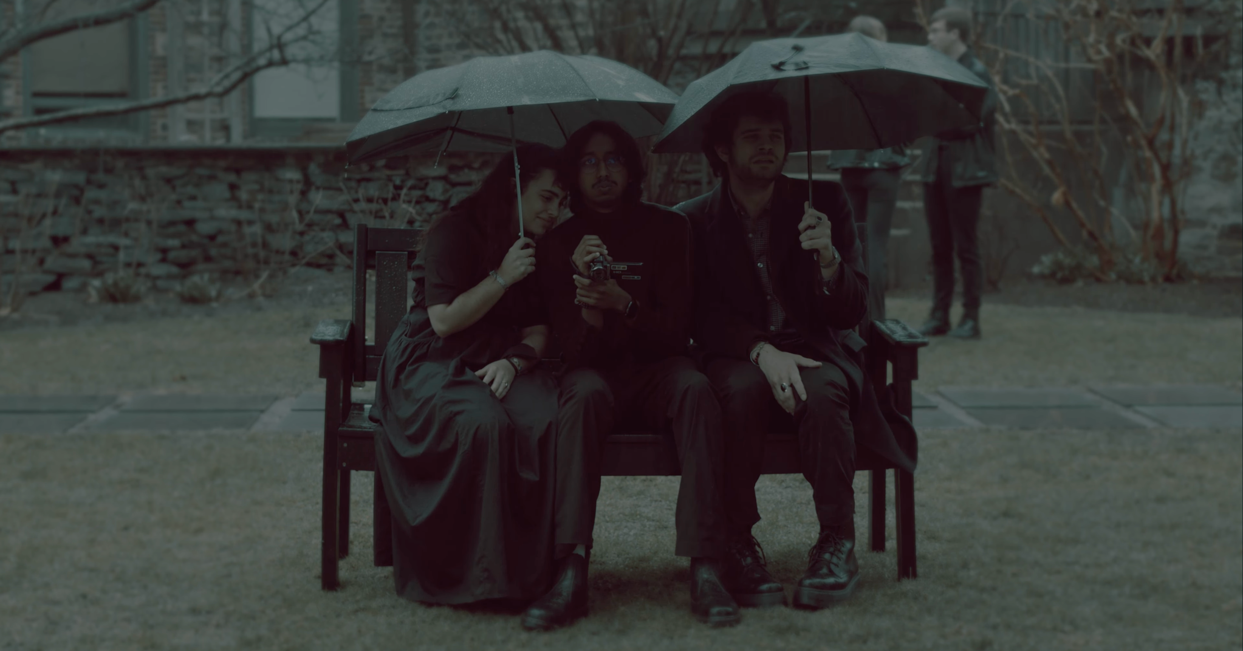 Three people sitting on a park bench holding umbrellas during rainy weather. One woman leaning on a man holding a camera, another man sitting beside them, all dressed in dark clothing. There are trees and a stone staircase in the background.