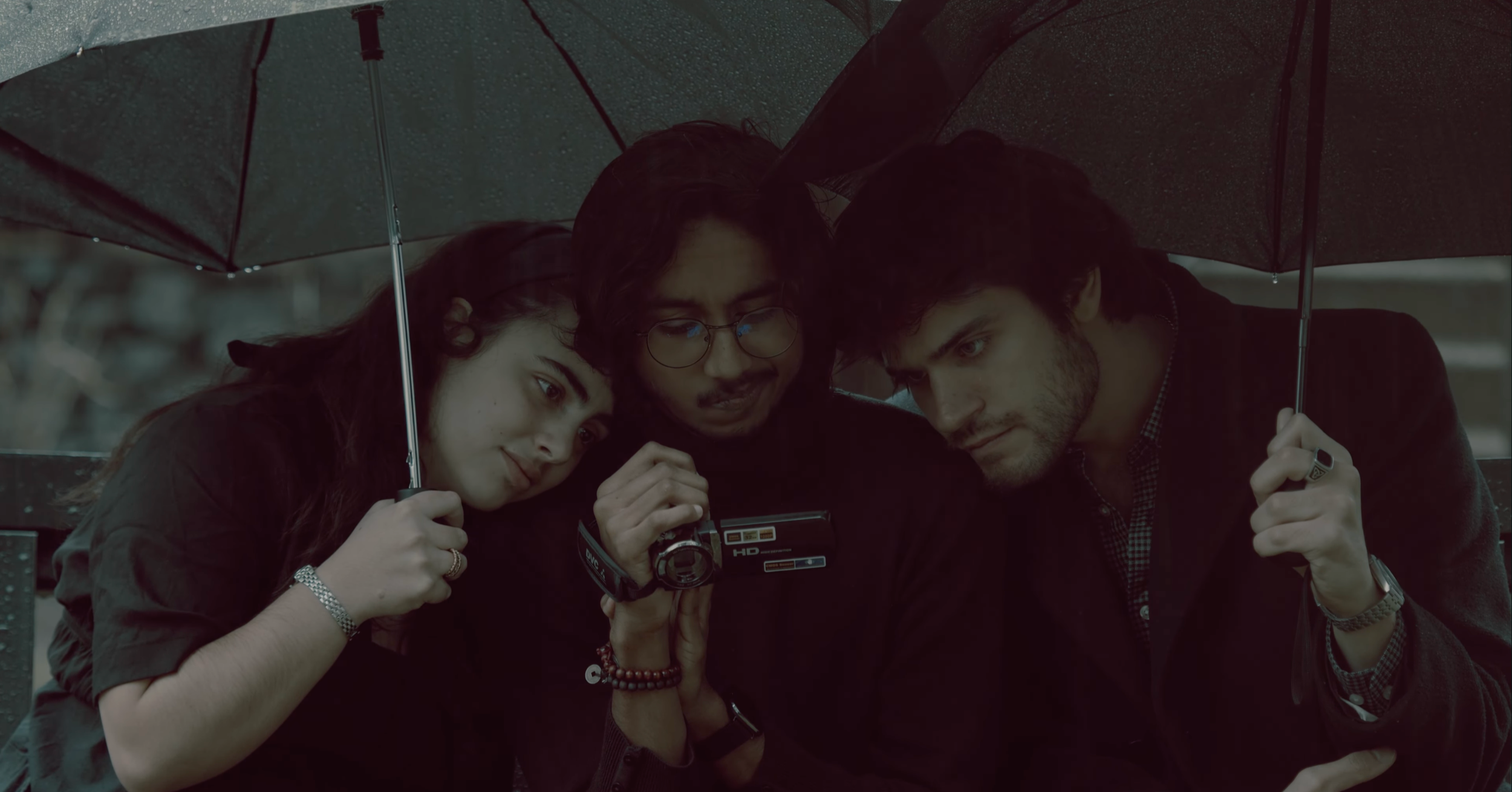 Three young people sitting closely together under a black umbrella in the rain, looking at a camera.