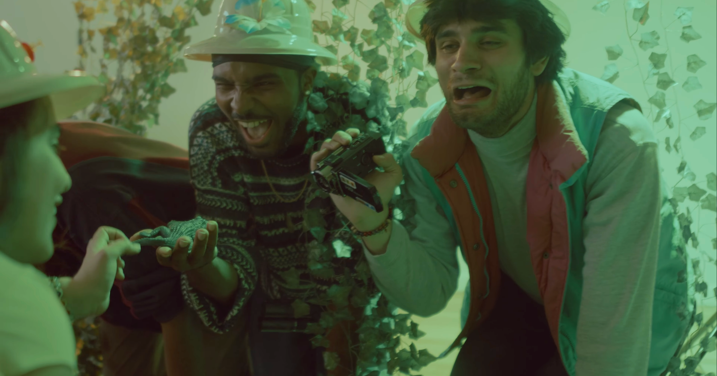 Young men in a green-lit room, one holding a camera and another with an expressive face, surrounded by plants and ivy.