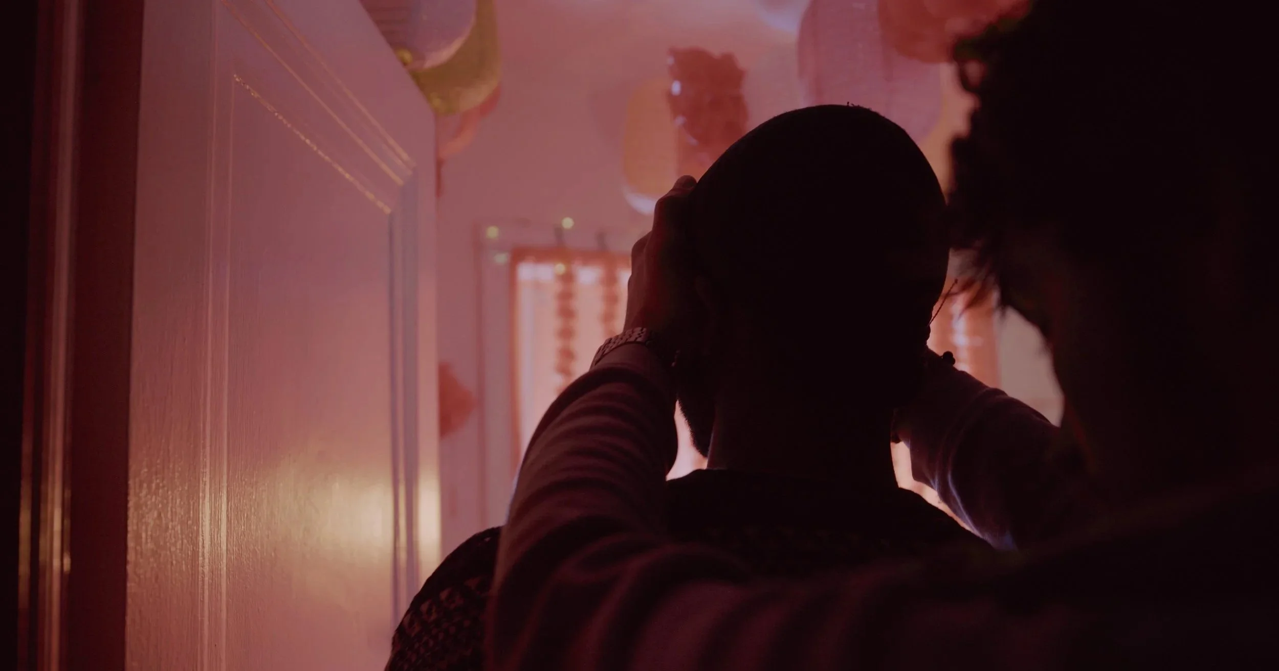 Person with curly hair and striped shirt, resting against a wall, silhouetted in warm, pinkish lighting with window blinds in the background.