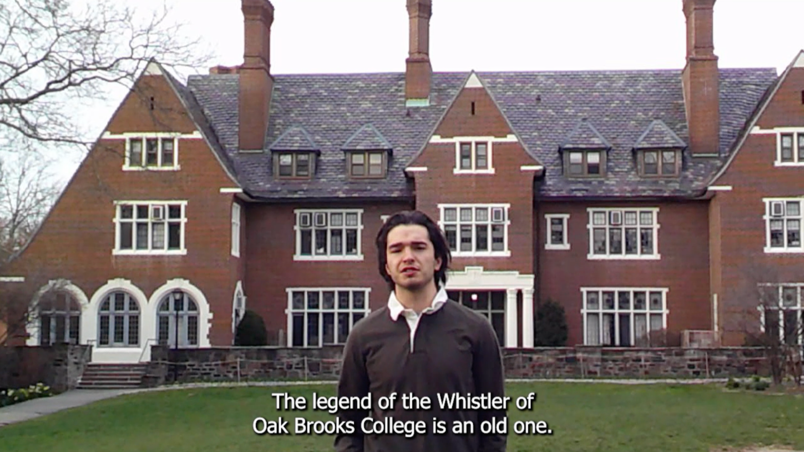 A young man standing in front of a large, historic brick mansion with multiple chimneys, pitched roofs, and numerous windows. The house is situated on a well-maintained lawn with a stone pathway and steps leading to the entrance.
