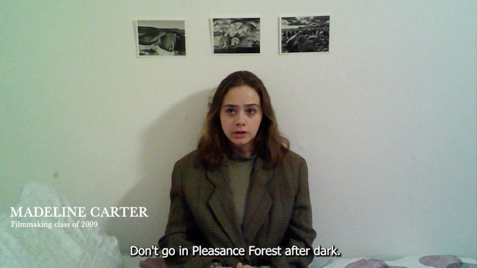 A young woman with shoulder-length brown hair sitting on a bed against a white wall with three black and white photographs. She is wearing a plaid blazer over a shirt. Subtitles read, "Don't go in Pleasance Forest after dark." Text in the bottom left