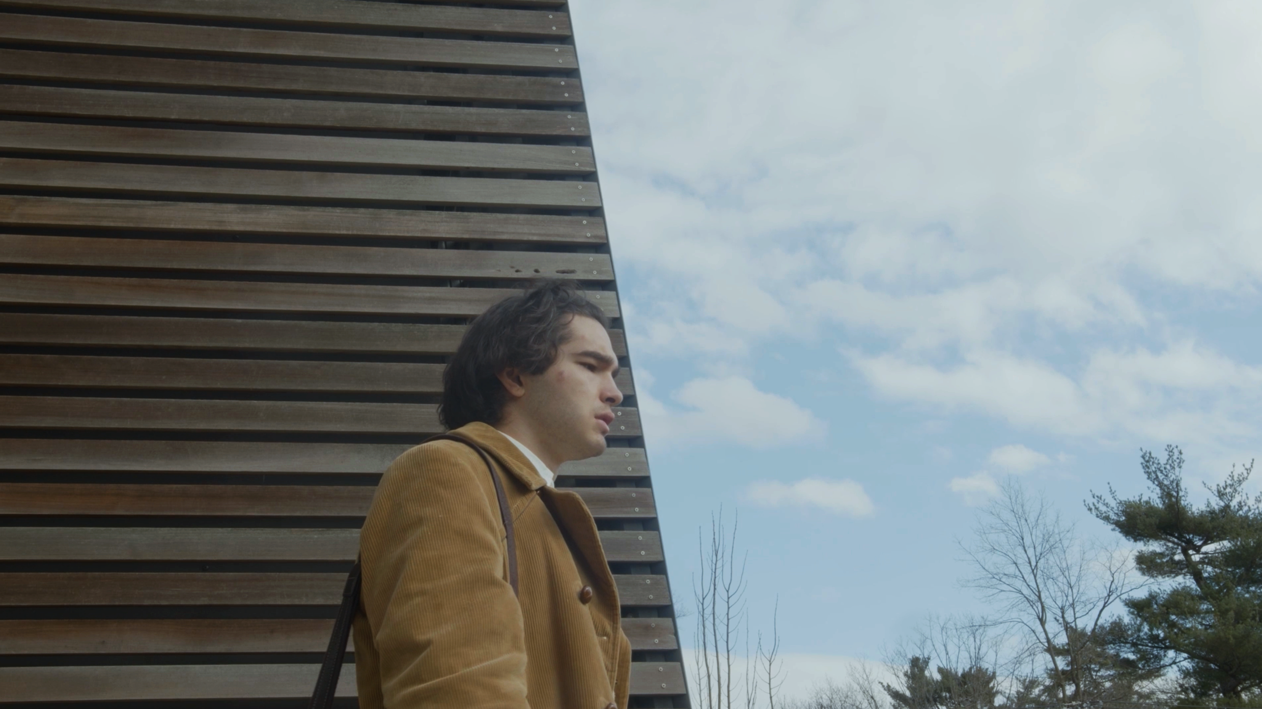 Young man with dark hair in a brown jacket standing outdoors near a wooden wall, with trees and clouds in the background.