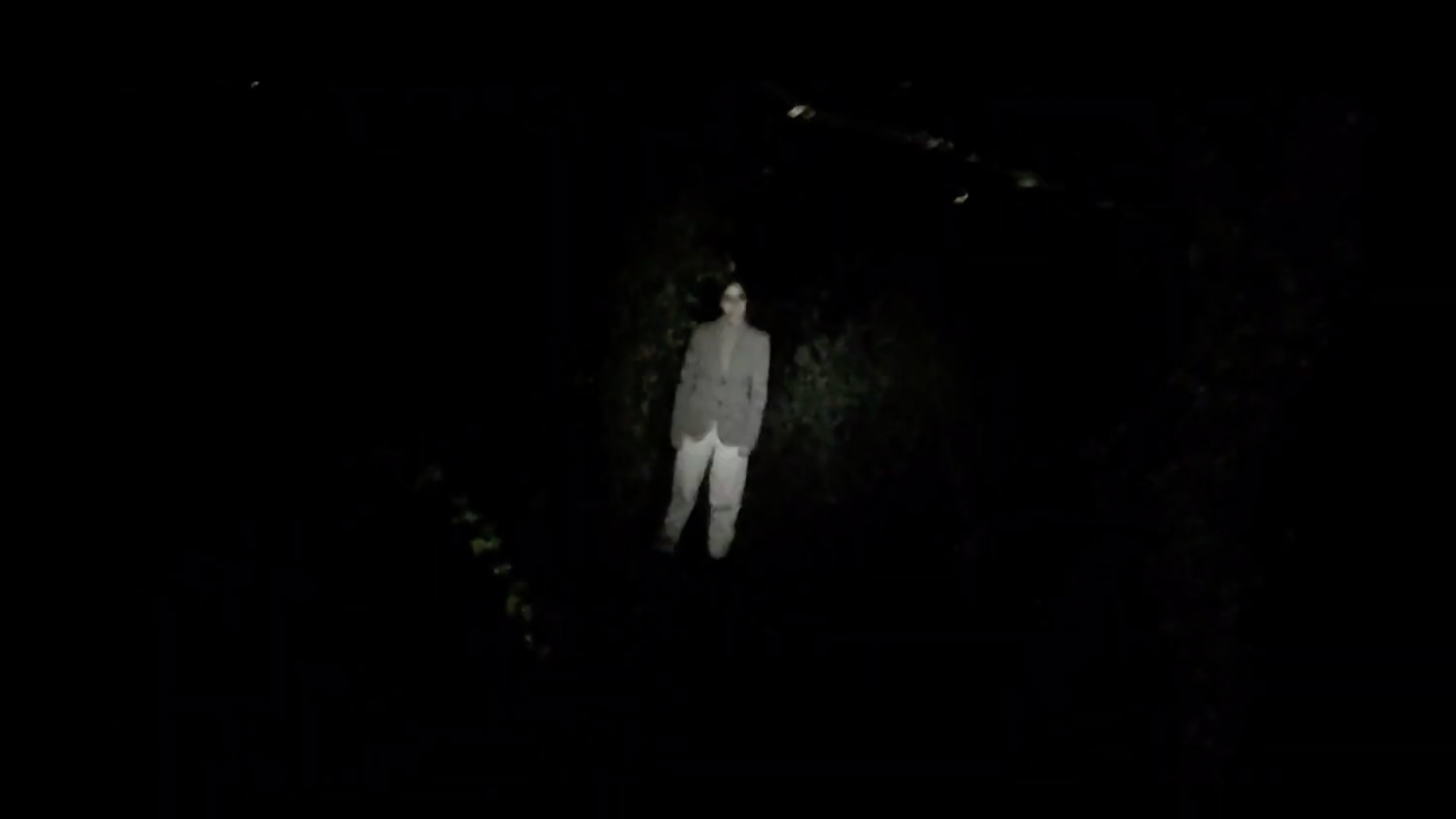 A woman wearing sunglasses and a blazer stands outdoors at night in front of dark trees.