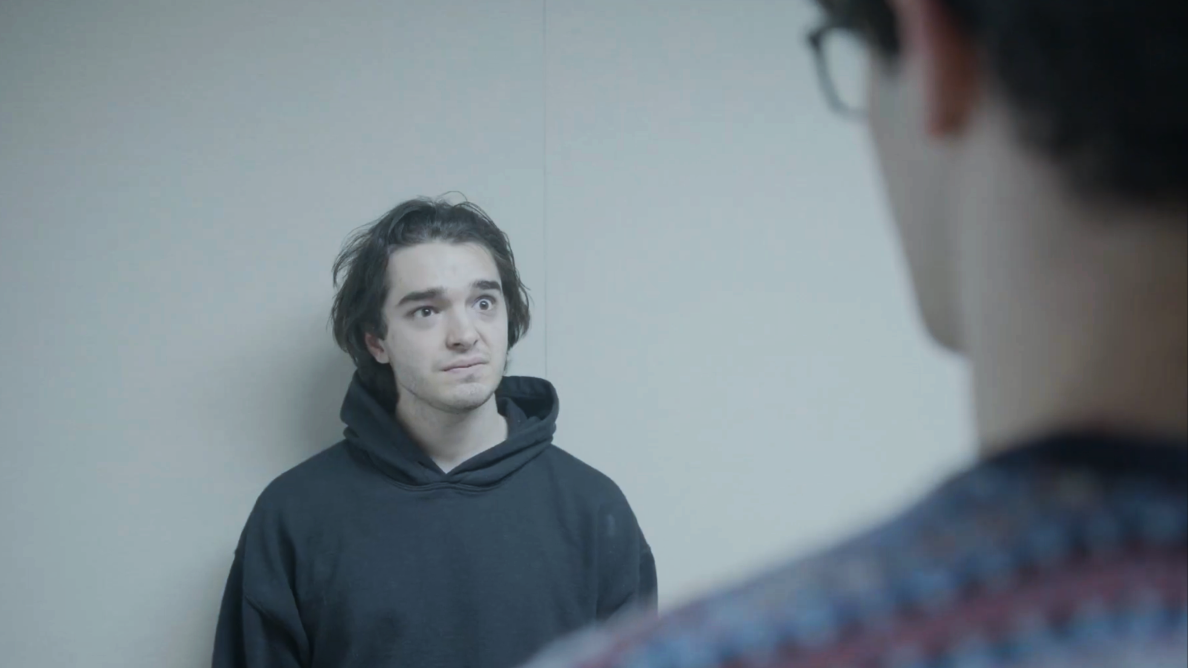 Image shows a young man with dark hair and a hoodie looking at his reflection in a mirror, with an out-of-focus person in the foreground.
