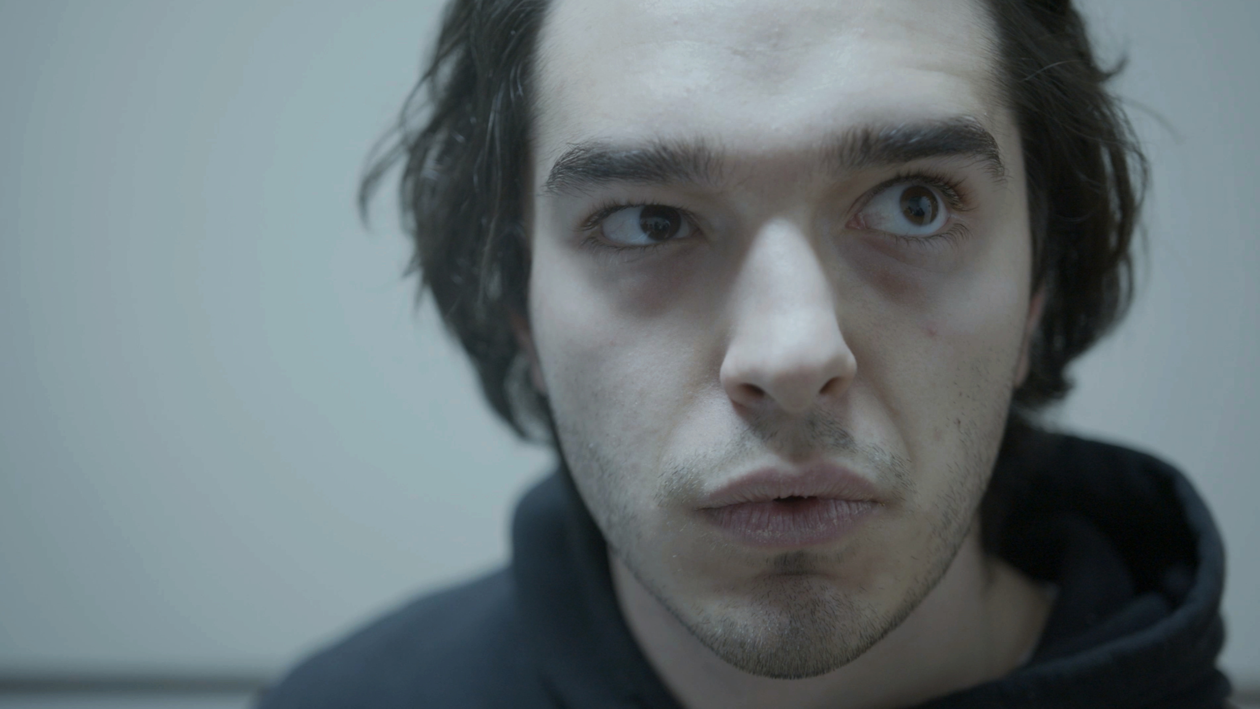 Close-up of a young man with dark, wavy hair and light skin, looking slightly upward and toward the camera, with a neutral expression against a plain, light gray background.