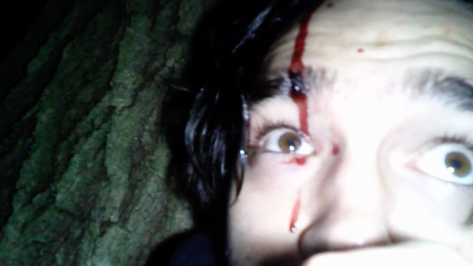 A woman with black hair lies on the ground with a bloody wound on her forehead, staring with wide eyes.