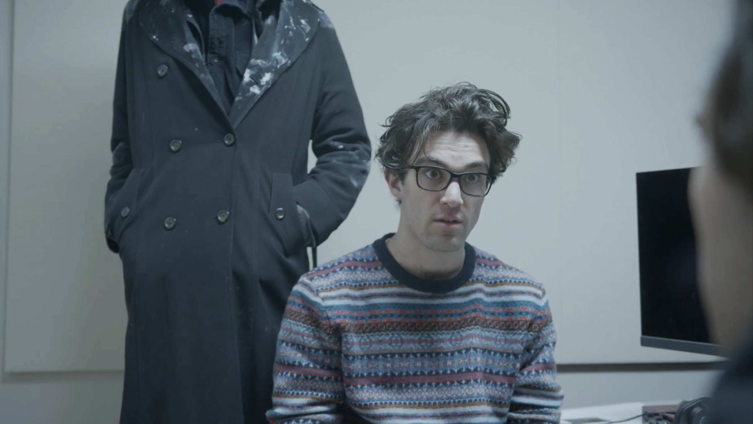 A man with dark, curly hair and glasses sitting at a desk looking at a computer monitor. A person in a dark coat standing next to him, with their hand in their pocket.