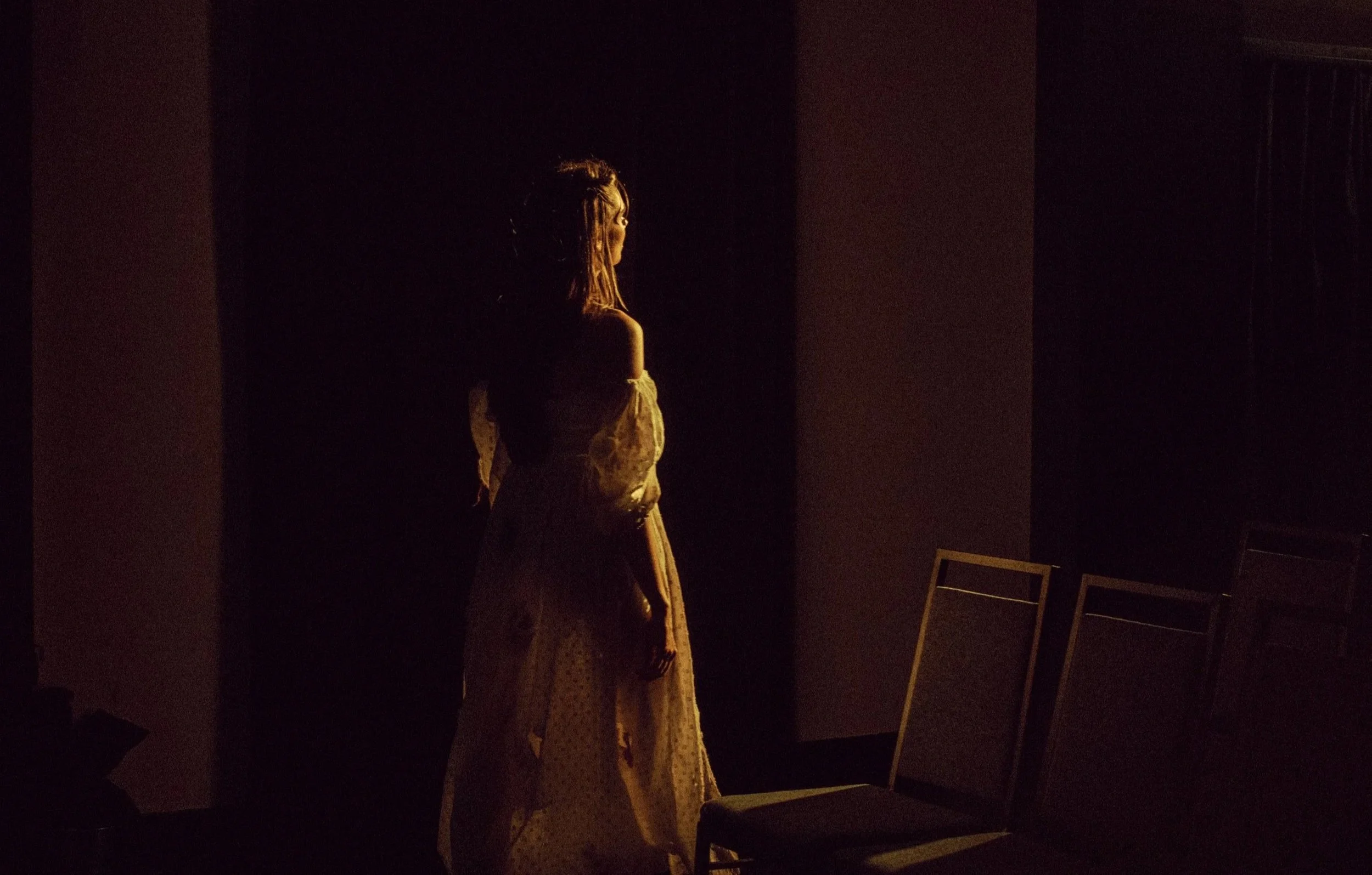 A woman standing in a dark room, illuminated softly from the side, wearing a light-colored, textured coat or dress with long sleeves.