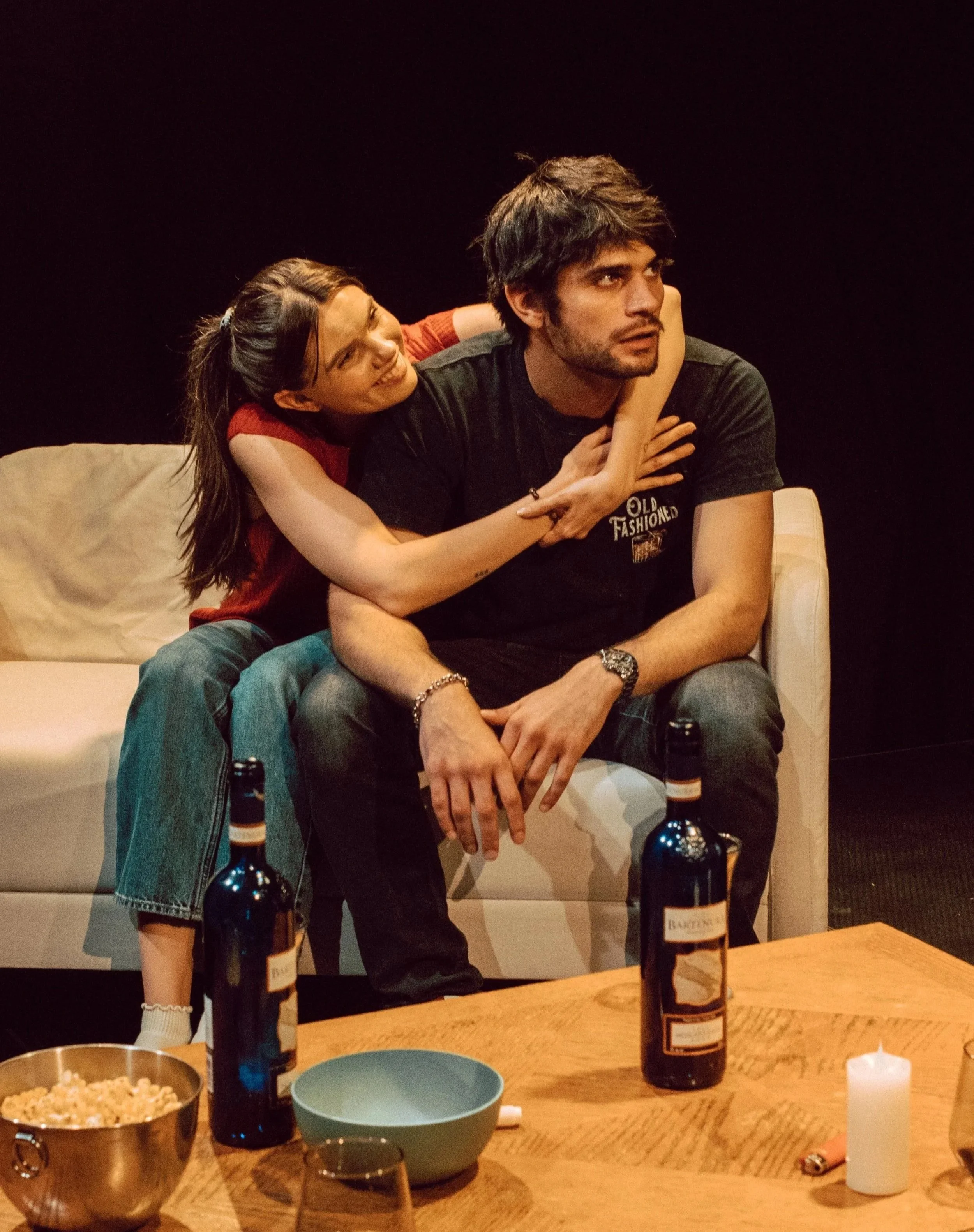 A woman with long dark hair and a young man with messy dark hair sitting closely on a beige couch, with the woman hugging the man from behind. The woman is smiling and looking at the man, who is looking away with a serious expression. On a wooden tab