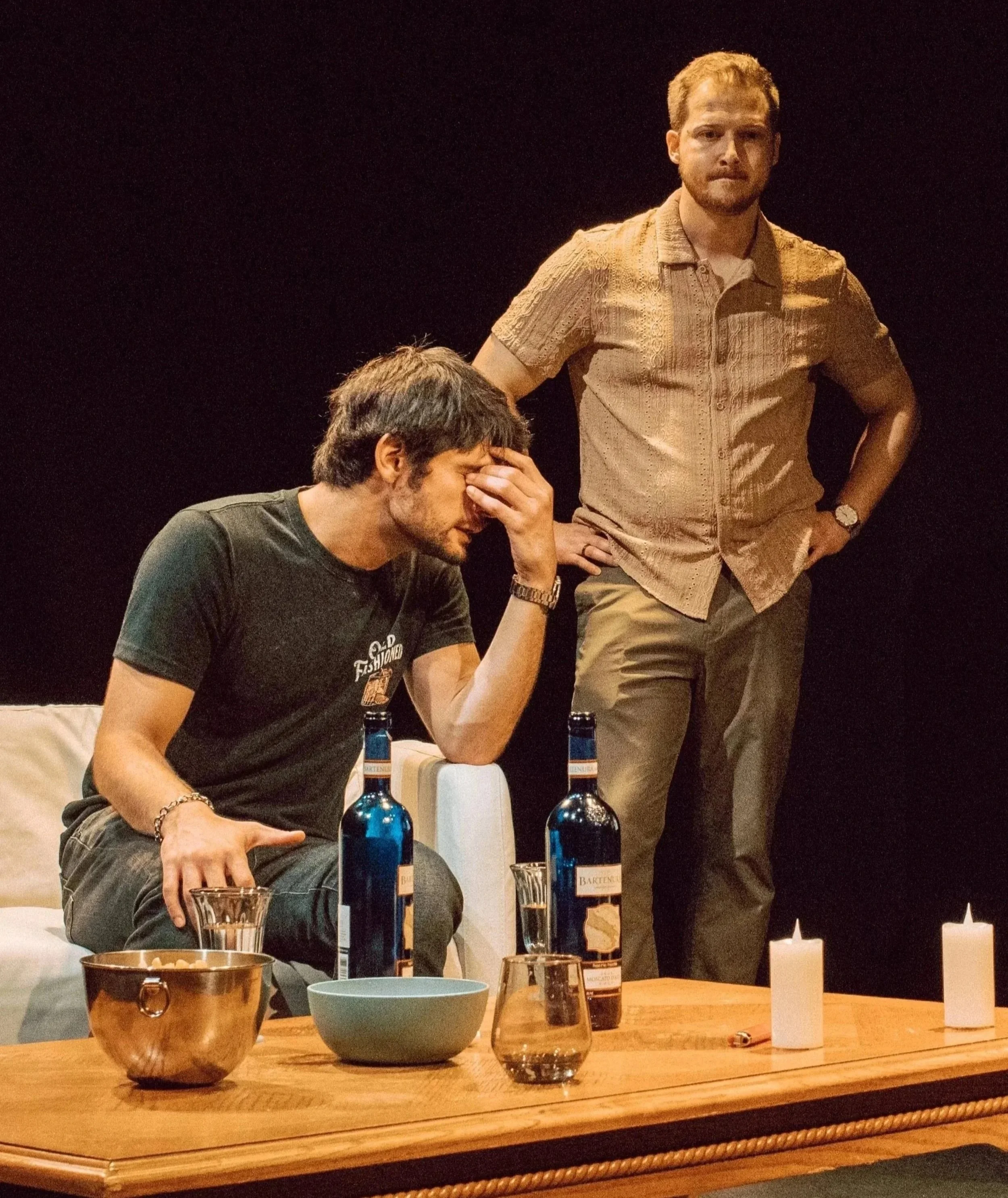Two men with drinks sitting at a table in a dimly lit room, and another standing man behind them. The table has bottles, a copper ice bucket, a bowl, and candles.