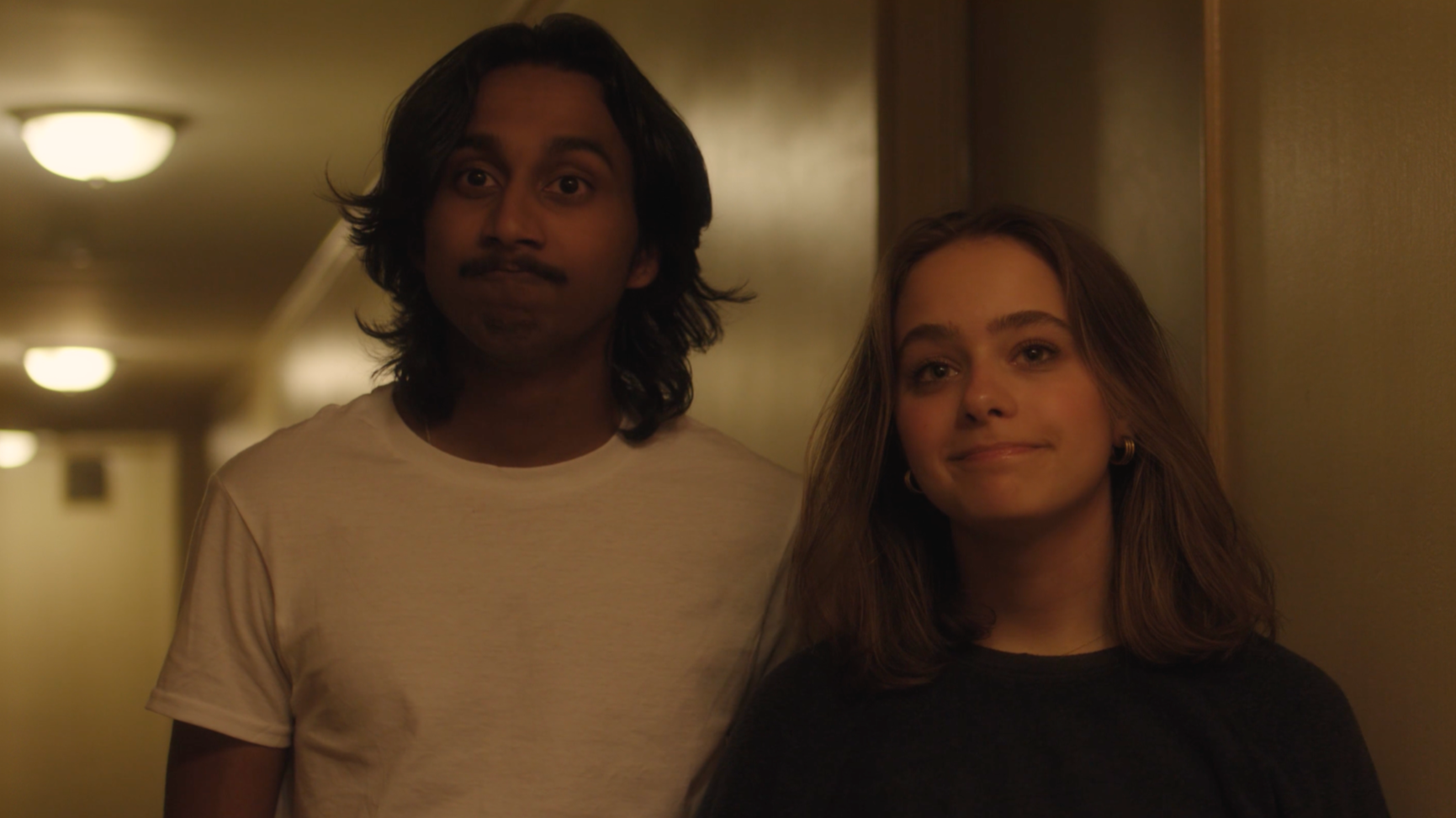 A scene from a movie showing a young man and woman standing in a dimly lit hallway with warm lighting and ceiling lights, both looking at the camera with neutral expressions.