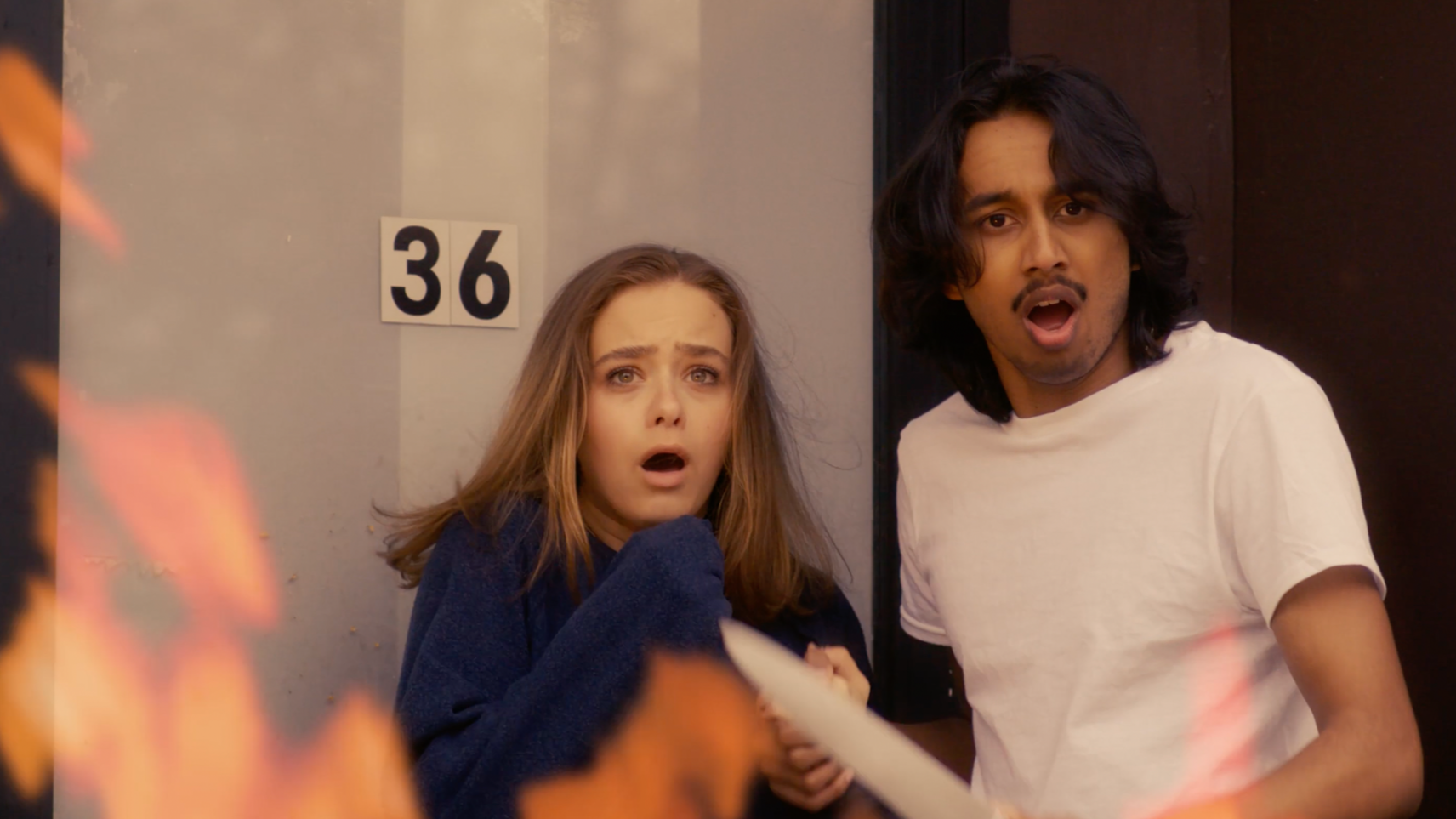 A woman and a man with shocked and scared expressions, standing in front of a wall with the number 36 on it. The woman has long brown hair and is wearing a blue sweater, while the man has shoulder-length black hair and is wearing a white t-shirt.