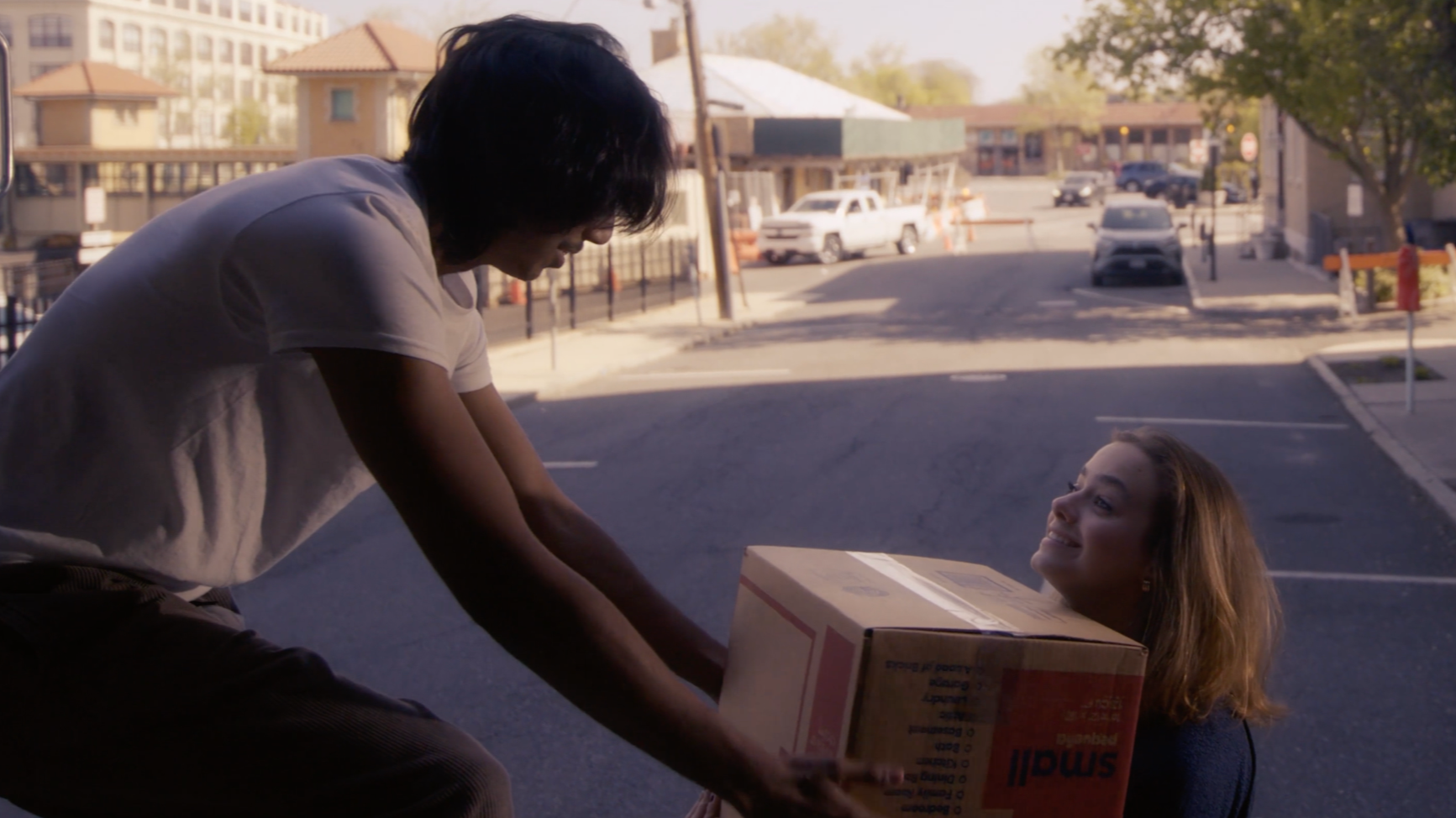 A person is giving a box to a woman sitting on the ground outside during daytime, with a parking lot and buildings in the background.