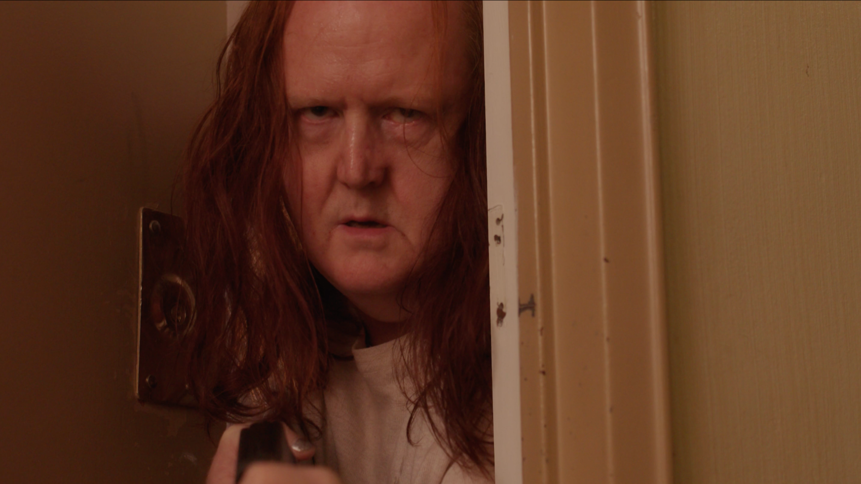 A person with long red hair and pale skin peering through a partially opened door, holding a key or similar object in their hand.