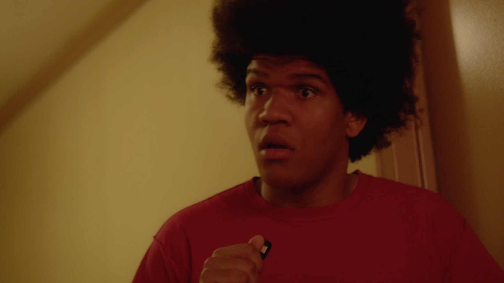 A man with a surprised or confused expression, holding a small object, standing in a doorway with warm-colored walls.