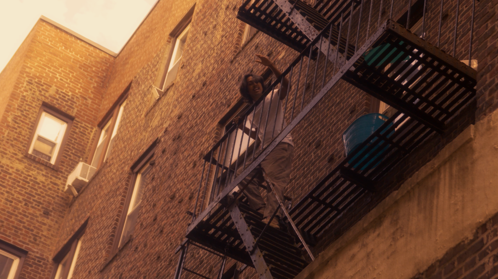A woman with dark hair, wearing a white shirt and tan pants, standing on an outdoor fire escape of a brick building, waving and smiling.