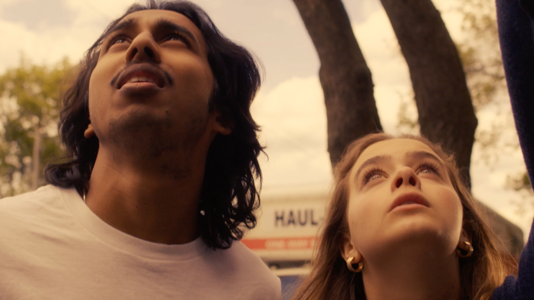 Close-up of a man with long dark hair and a woman with long light brown hair, both looking upward outdoors during daytime.