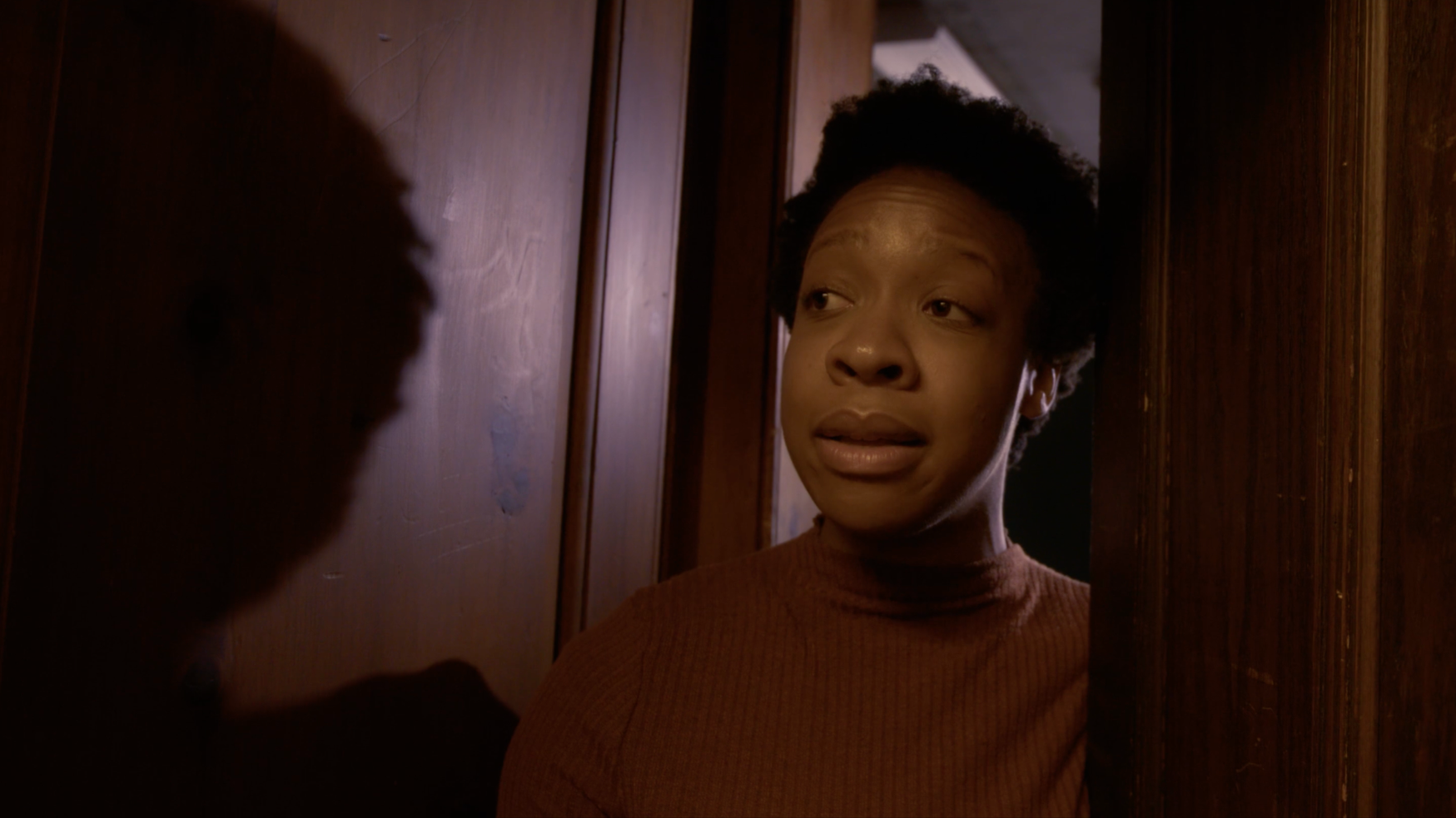 A woman with short curly hair and a brown sweater is speaking to a person, partially visible, through a wooden door or wall.
