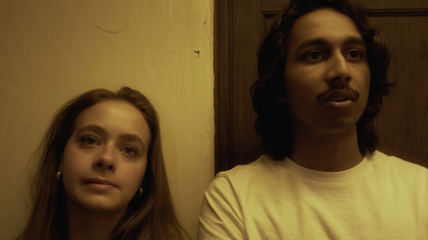 A woman with long brown hair and earrings, and a man with shoulder-length dark hair and a mustache, standing against a beige wall with a wooden door in the background.