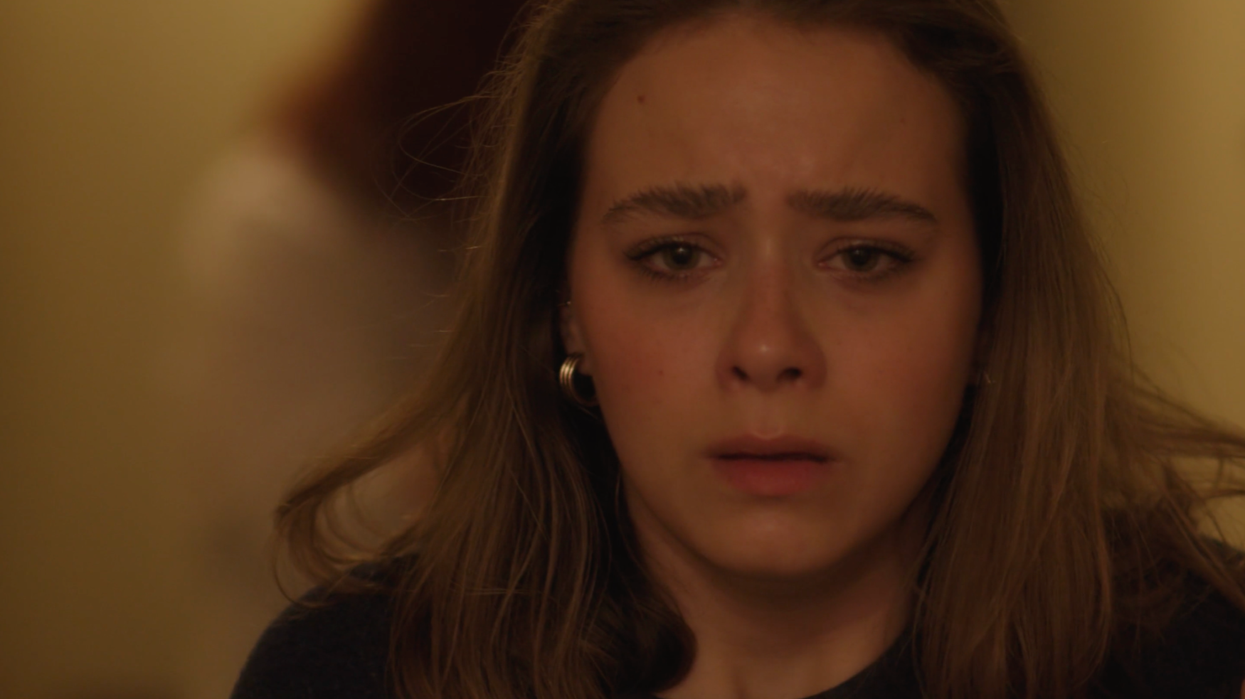 A young woman with light skin and long brown hair appears distressed, with a sad expression on her face.