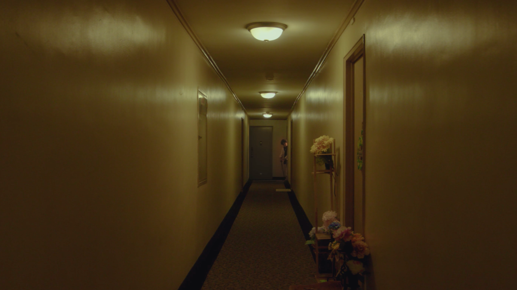 A dimly lit hotel hallway with closed doors on either side, floral arrangements on a small table, and a person standing at the end, partially obscured.