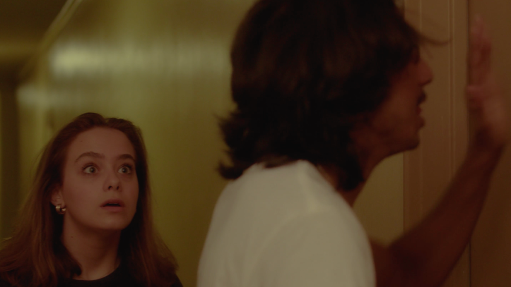 A young woman with long red hair, wearing earrings, looks surprised as she looks at a man with long dark hair, who is pressing his hand against a wall and facing away from her.