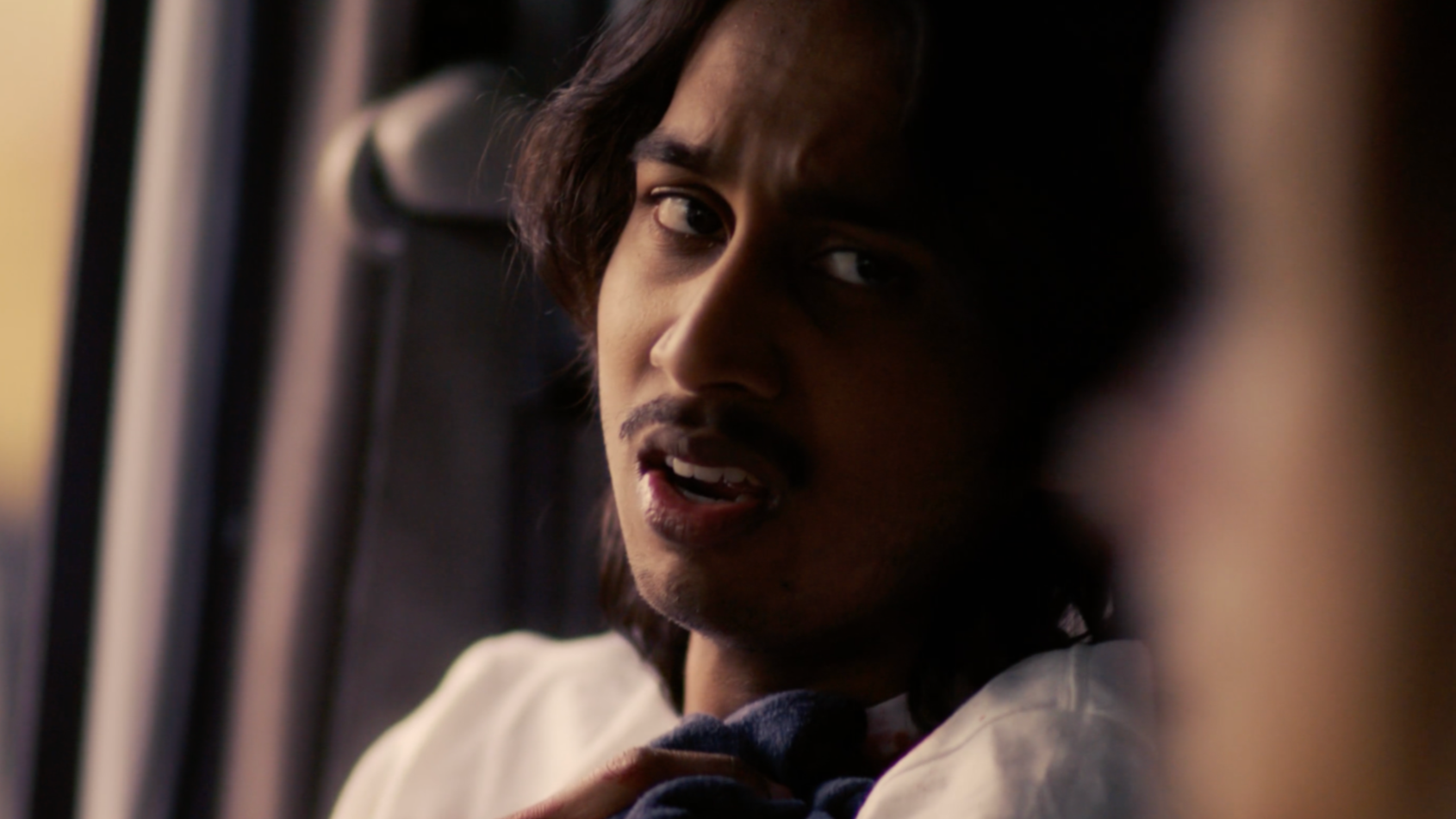A young man with shoulder-length hair and a mustache looks off to the side with a serious expression inside a vehicle, possibly a bus or train.